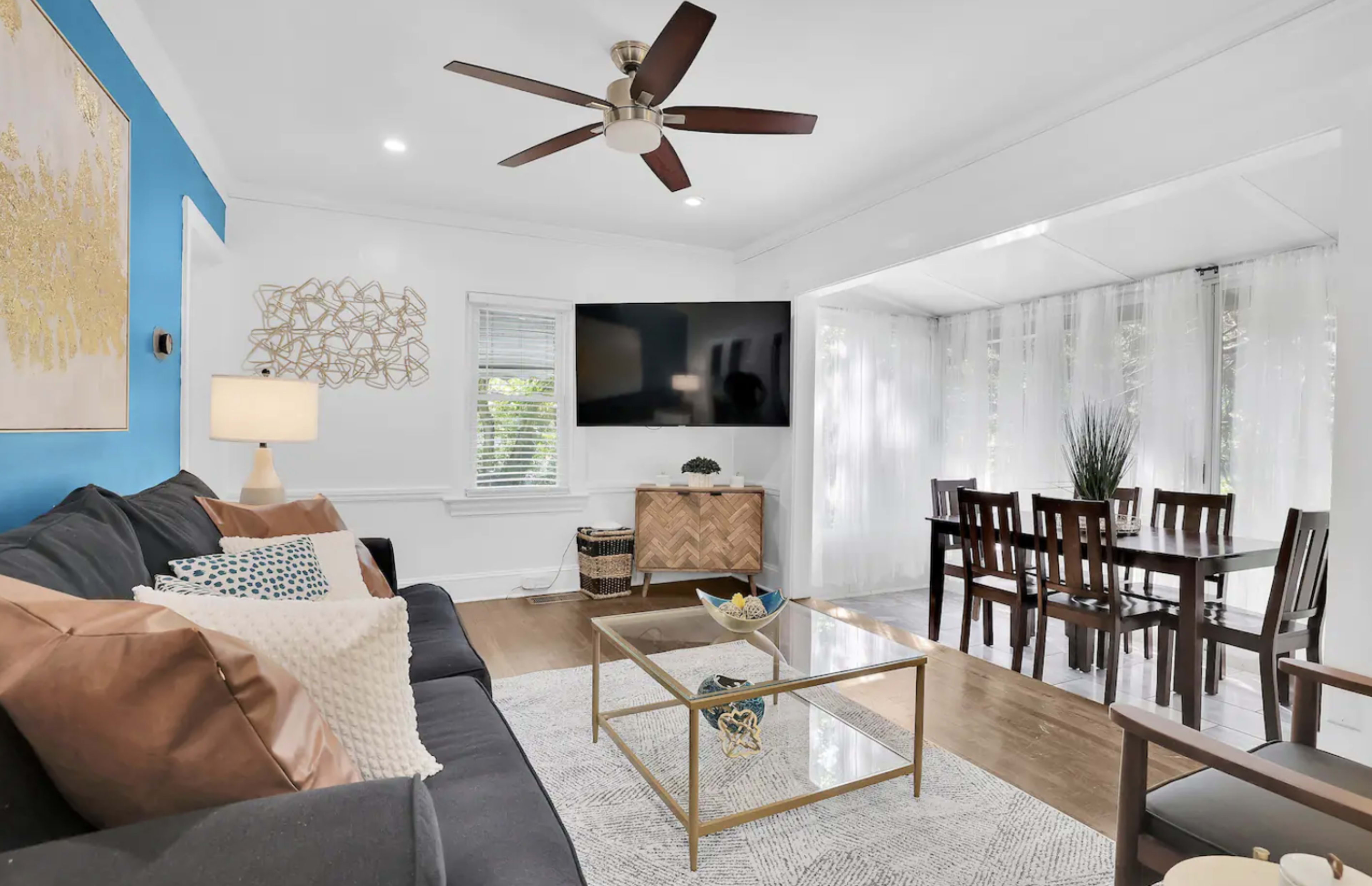 A living room features a dark sofa, a glass coffee table, and a wall-mounted television, with a dining area visible through a bright, airy space adorned with white curtains.