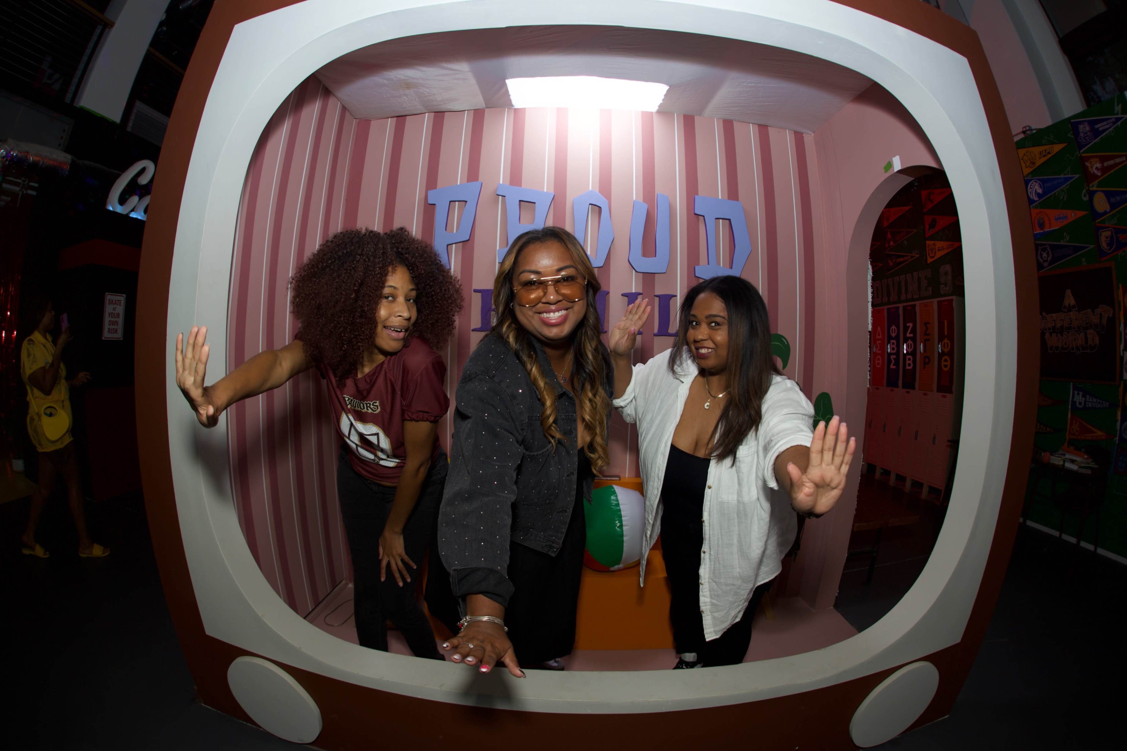 Three women are posing playfully inside a large, retro-styled photo booth with a colorful background.