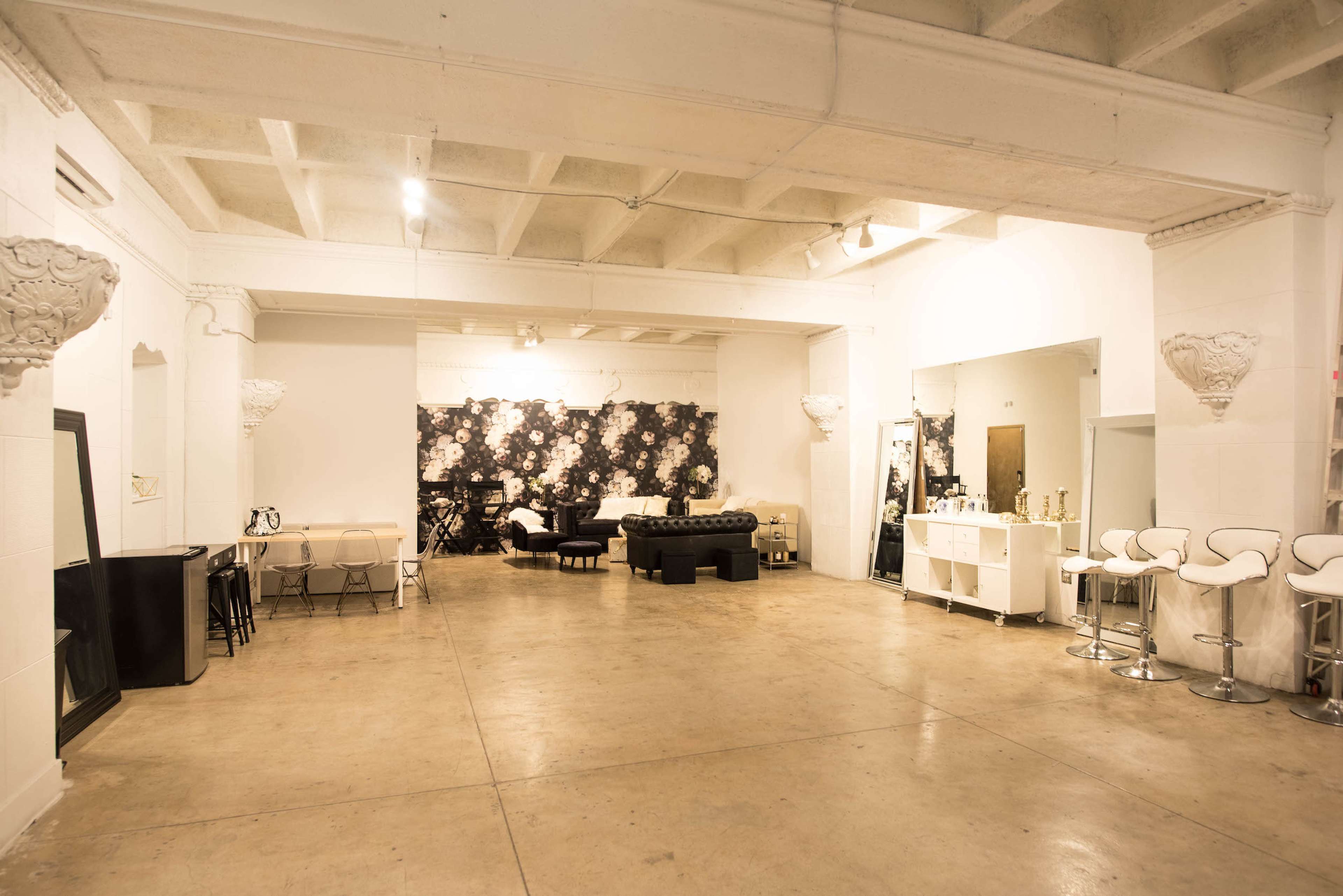 The image shows a spacious indoor area with a mix of seating arrangements including a black couch, white chairs, and a bar counter against a wall adorned with floral patterns.