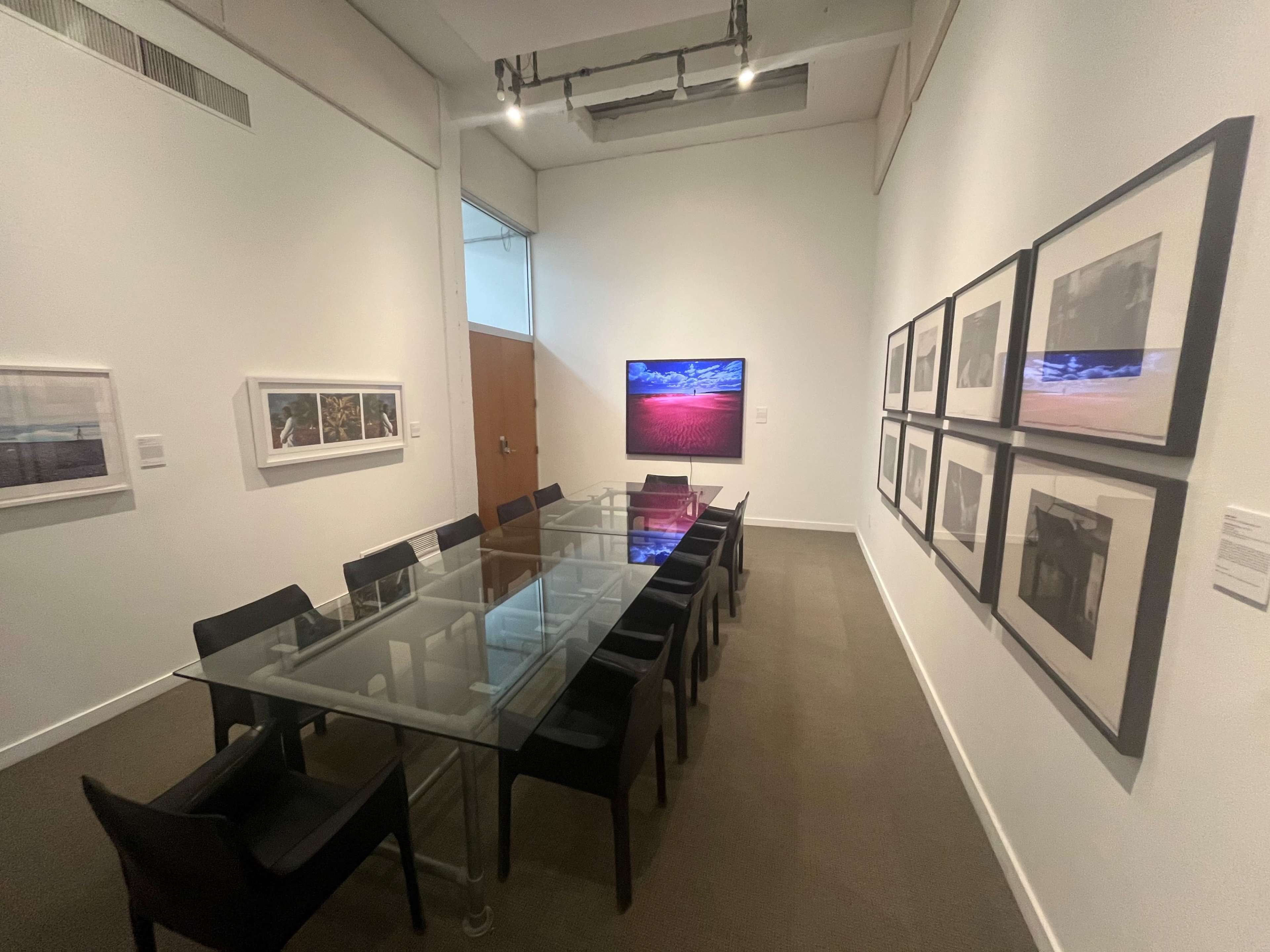 A long glass conference table is set with black chairs, surrounded by white walls displaying framed photographs in a well-lit room.