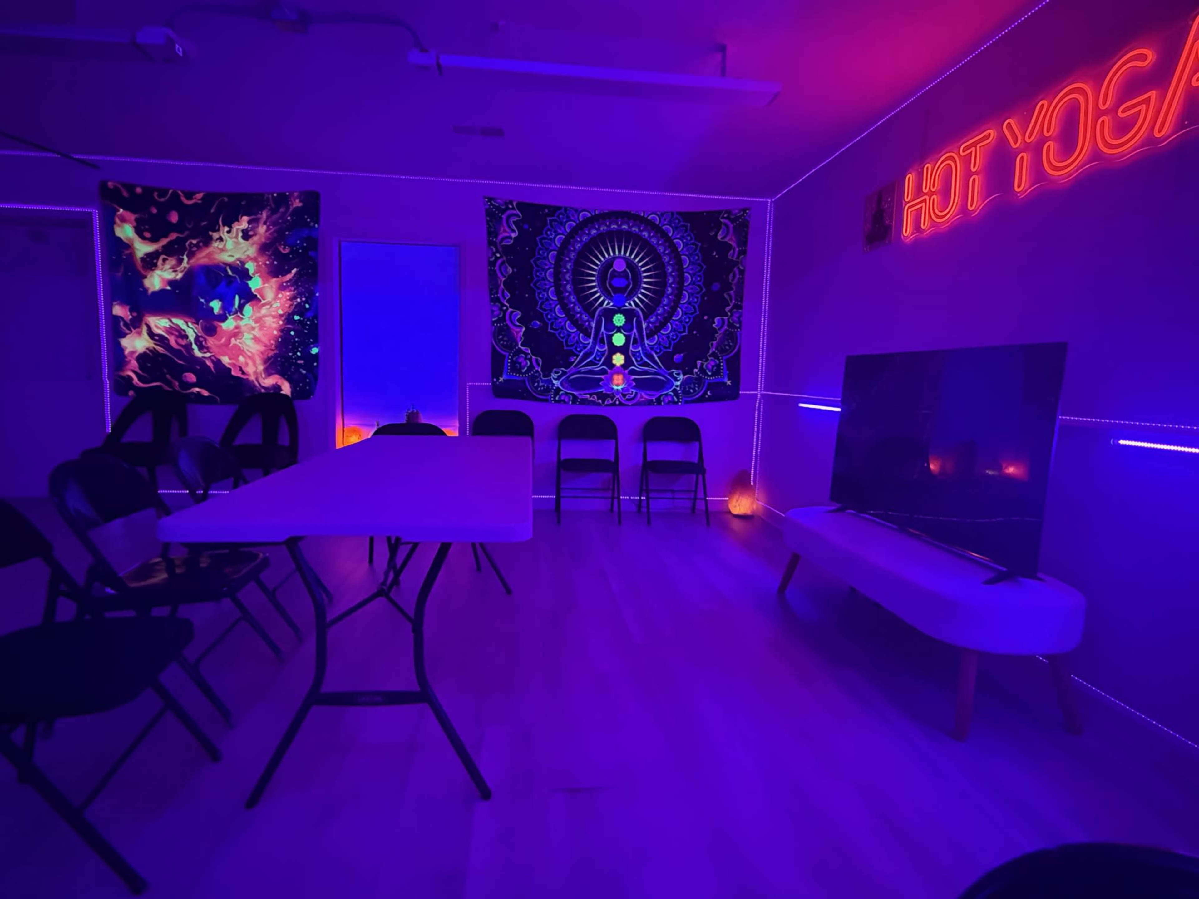 The image shows a dimly lit room designed for yoga, featuring a table, several chairs, colorful wall art, a television, and neon lighting.
