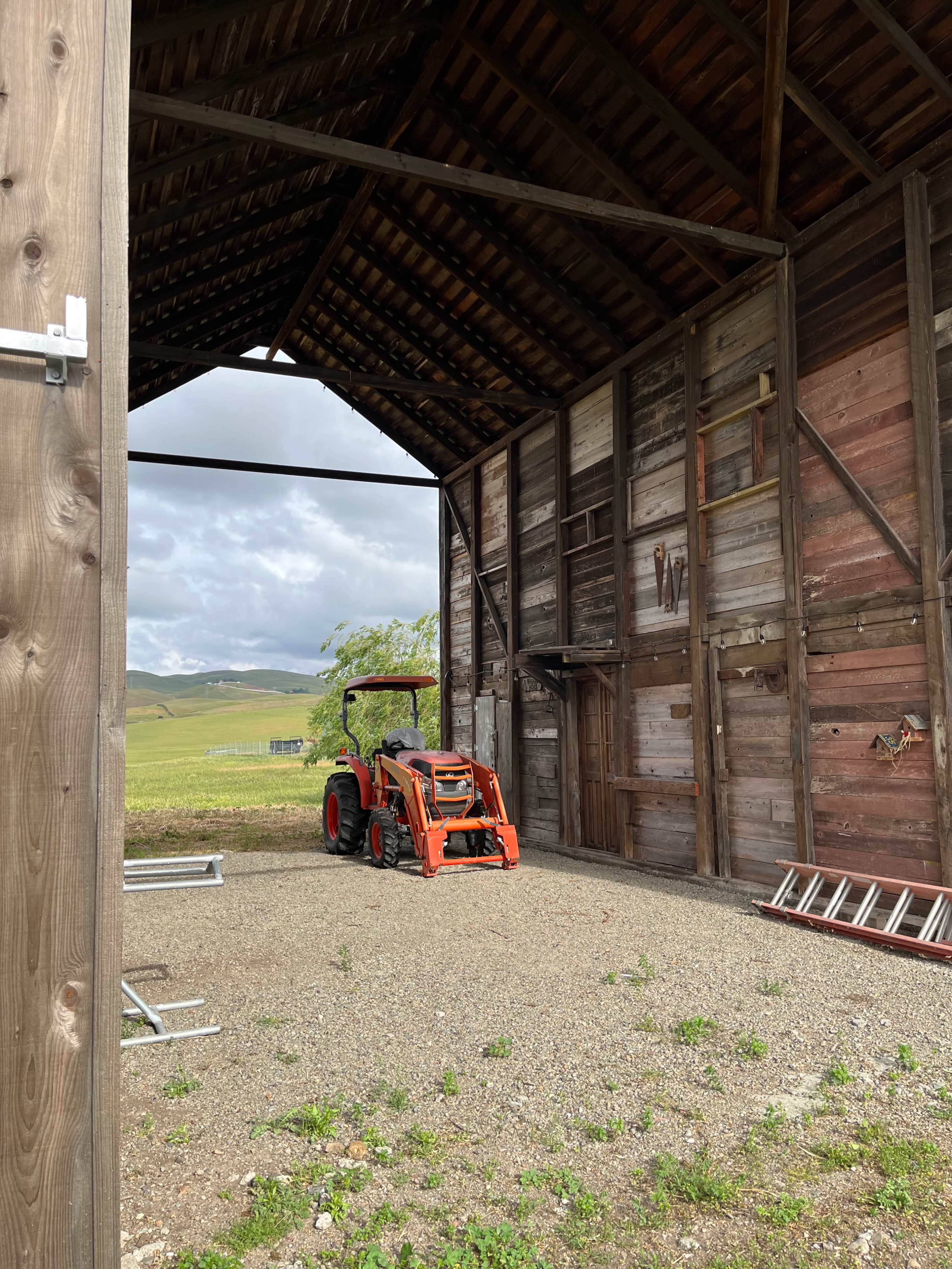 An orange tractor is parked inside a large, open barn with wooden walls and a gravel floor.