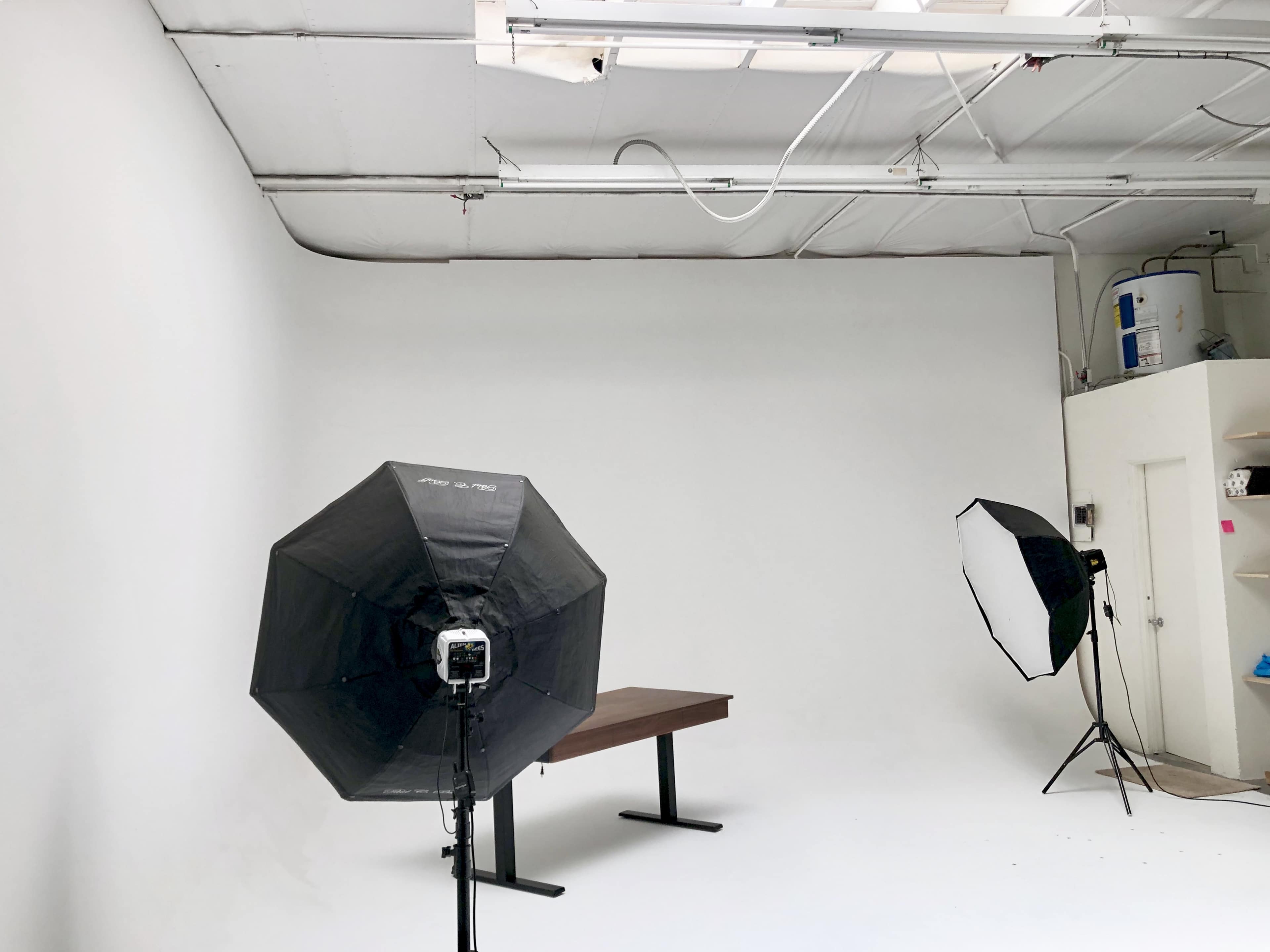 The image shows a photography studio with a plain white backdrop, a wooden table in the center, and two lighting setups positioned on either side.