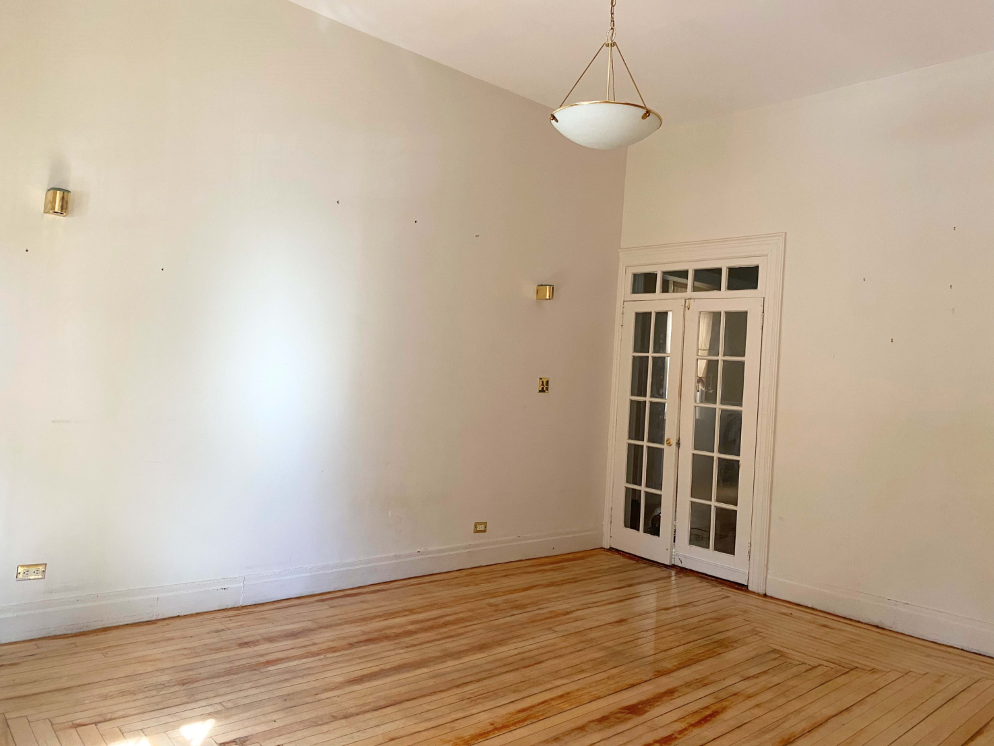 The image shows a spacious room with hardwood floors, white walls, and a set of double doors leading to another space, illuminated by a hanging light fixture.