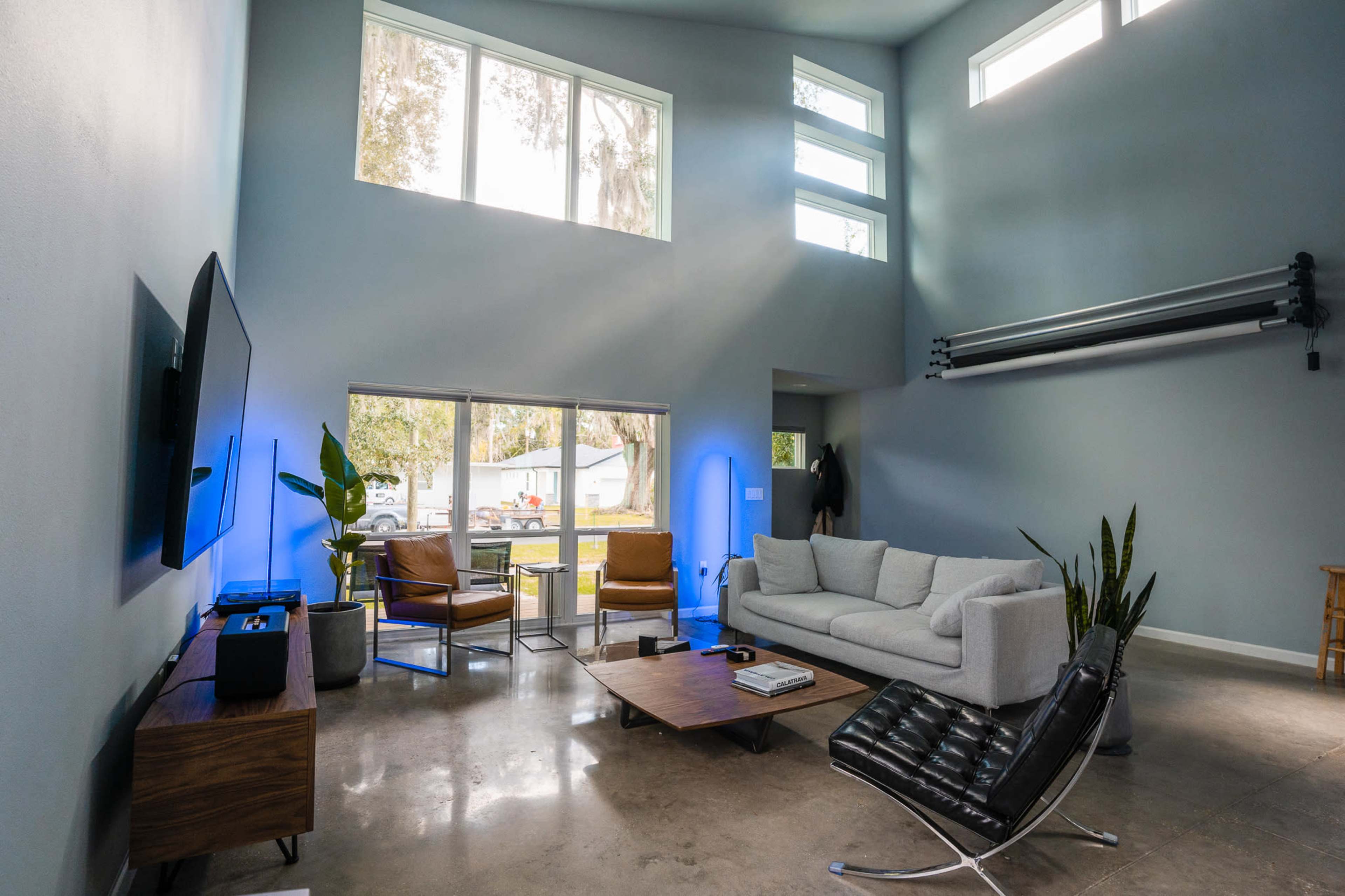 A modern living room with a gray sofa, a wooden coffee table, a television mounted on the wall, and large windows letting in natural light.