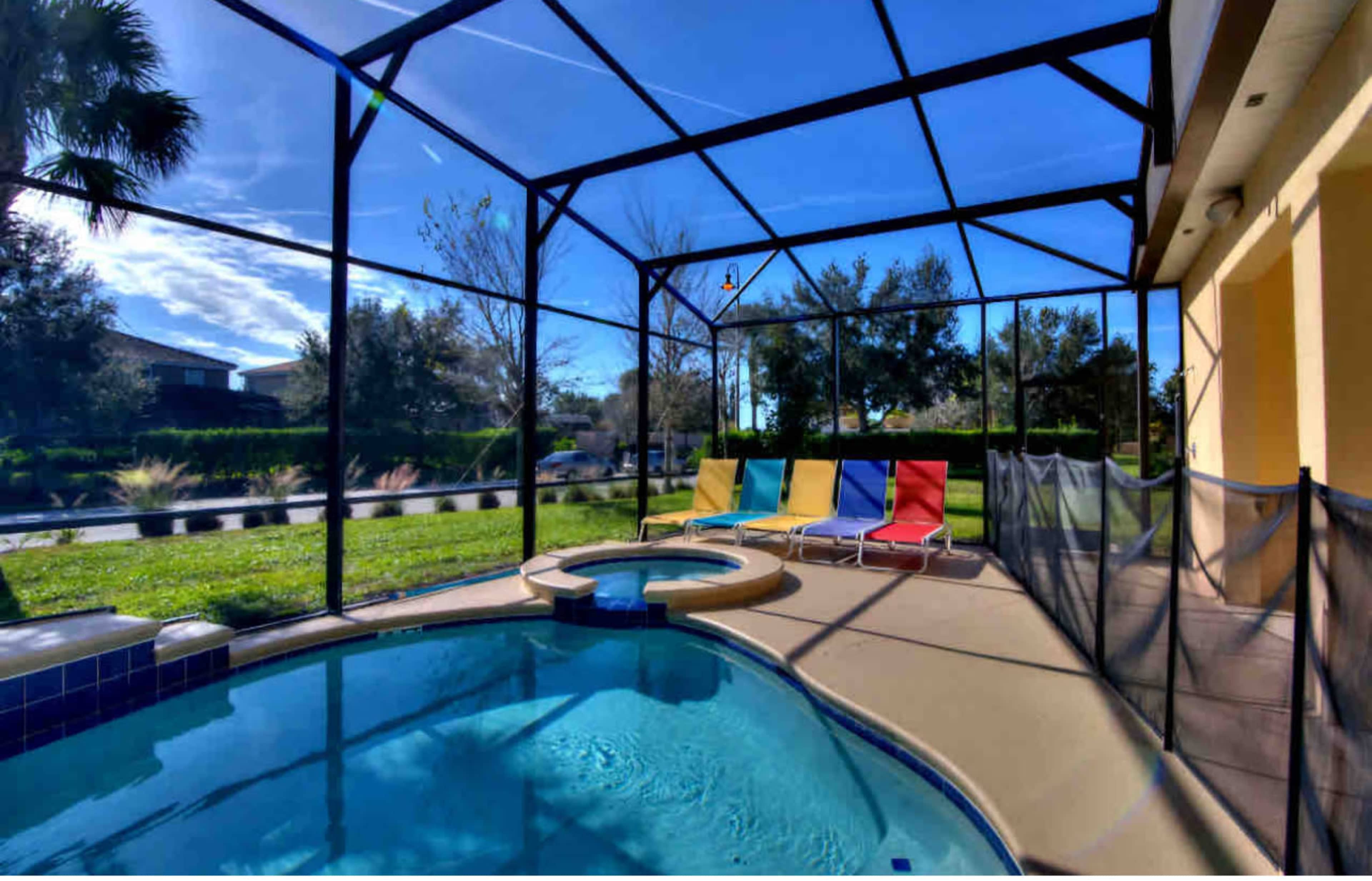 A screened-in pool area with a circular hot tub, colorful lounge chairs, and a view of a manicured lawn.