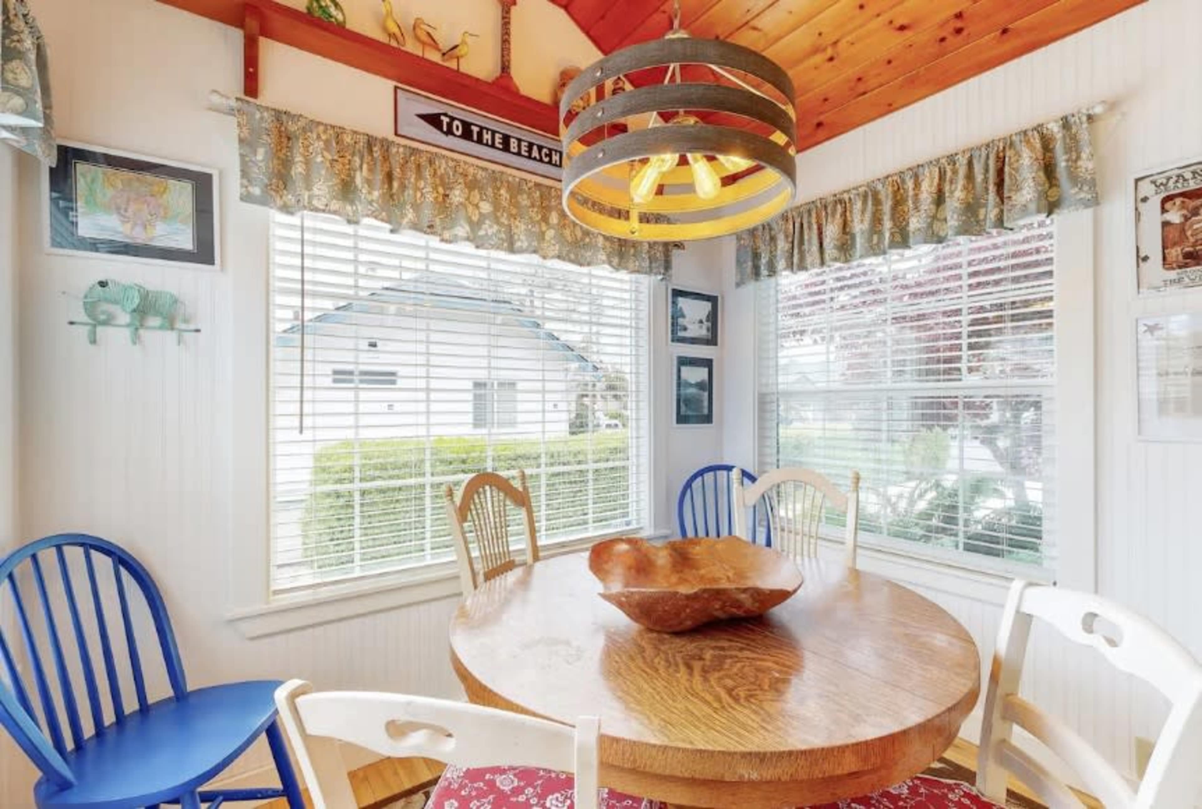 A round wooden table sits in a brightly lit dining area surrounded by four colorful chairs near a window with decorative curtains.