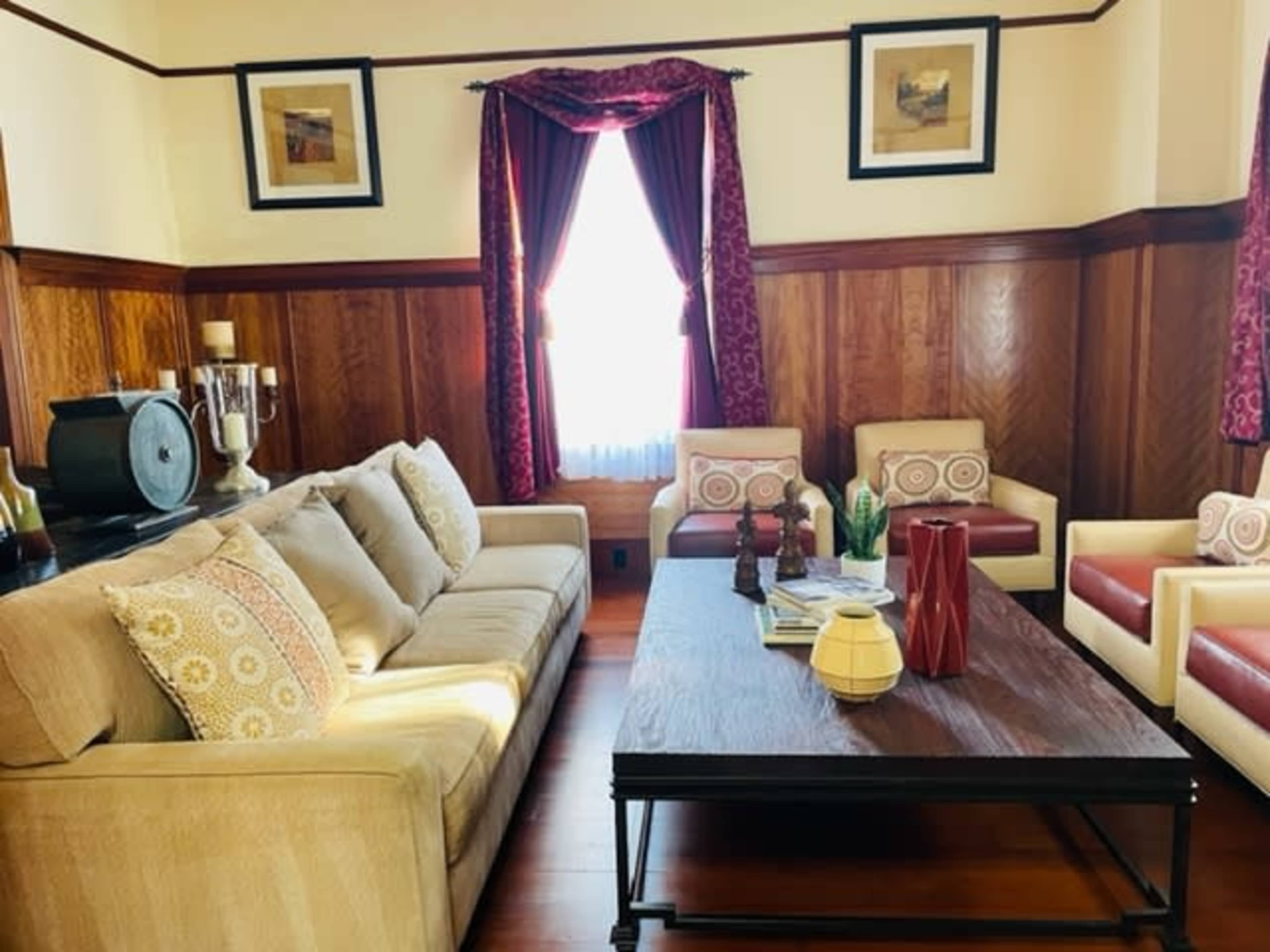 A living room with a beige sofa, a dark wooden coffee table, and two red and cream armchairs, all set against wooden-paneled walls and decorated with framed artwork.