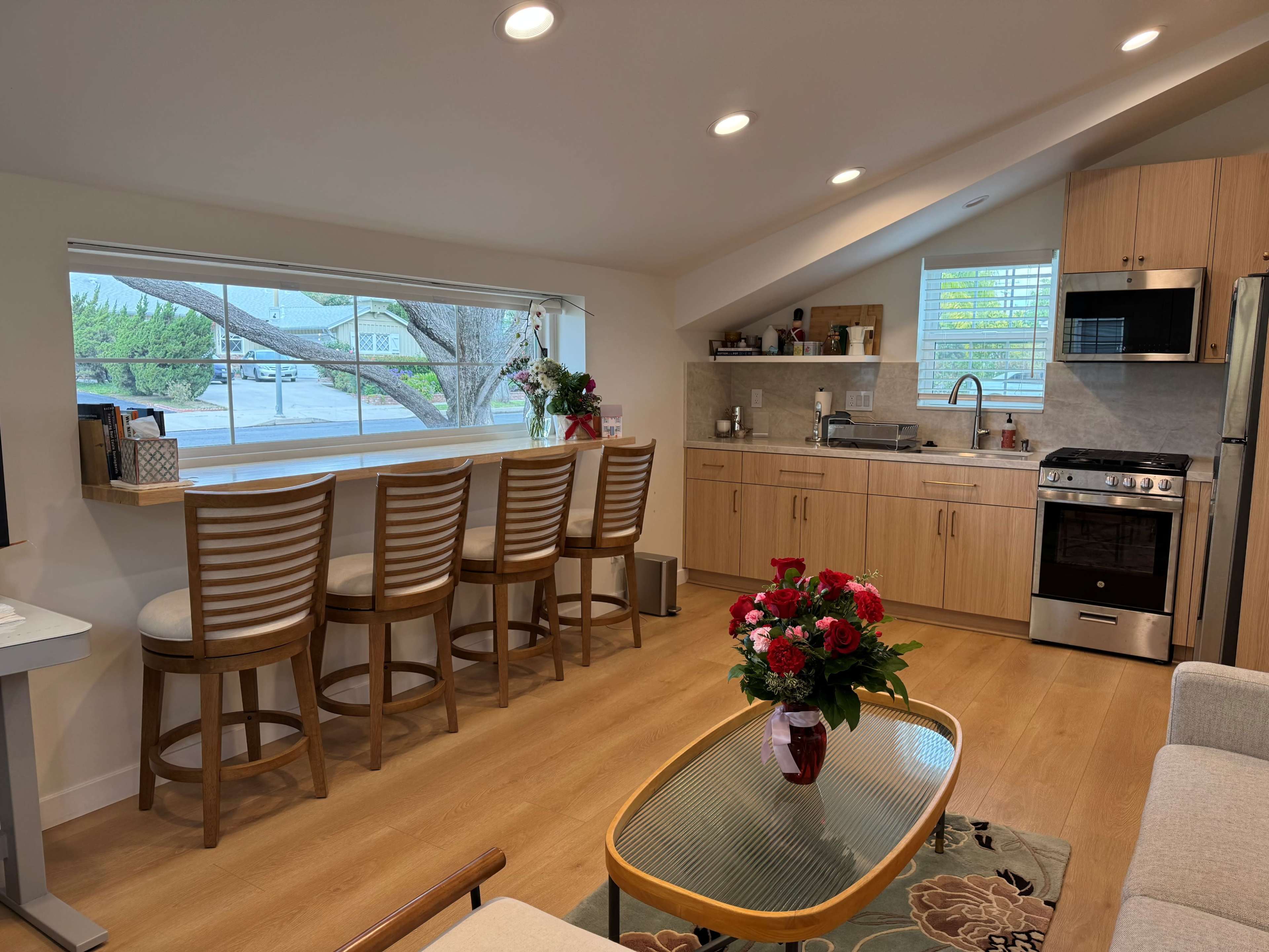 The image shows a modern kitchen with a breakfast bar and seating, featuring light wood cabinetry, stainless steel appliances, and a window that offers a view of the outdoors.