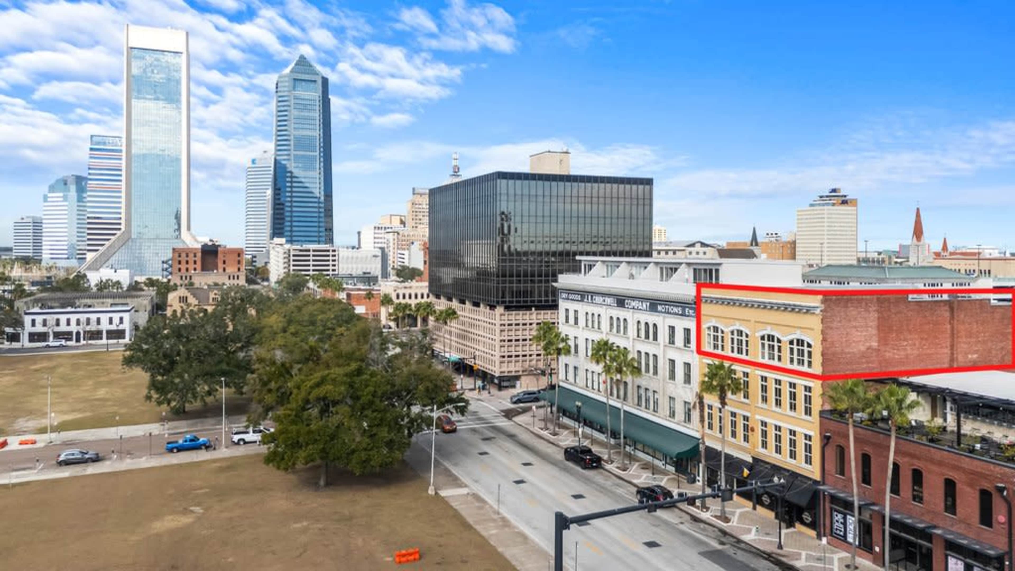 The image shows a cityscape of Jacksonville, Florida, featuring a mix of modern skyscrapers and historic buildings, with a street lined by palm trees in the foreground.