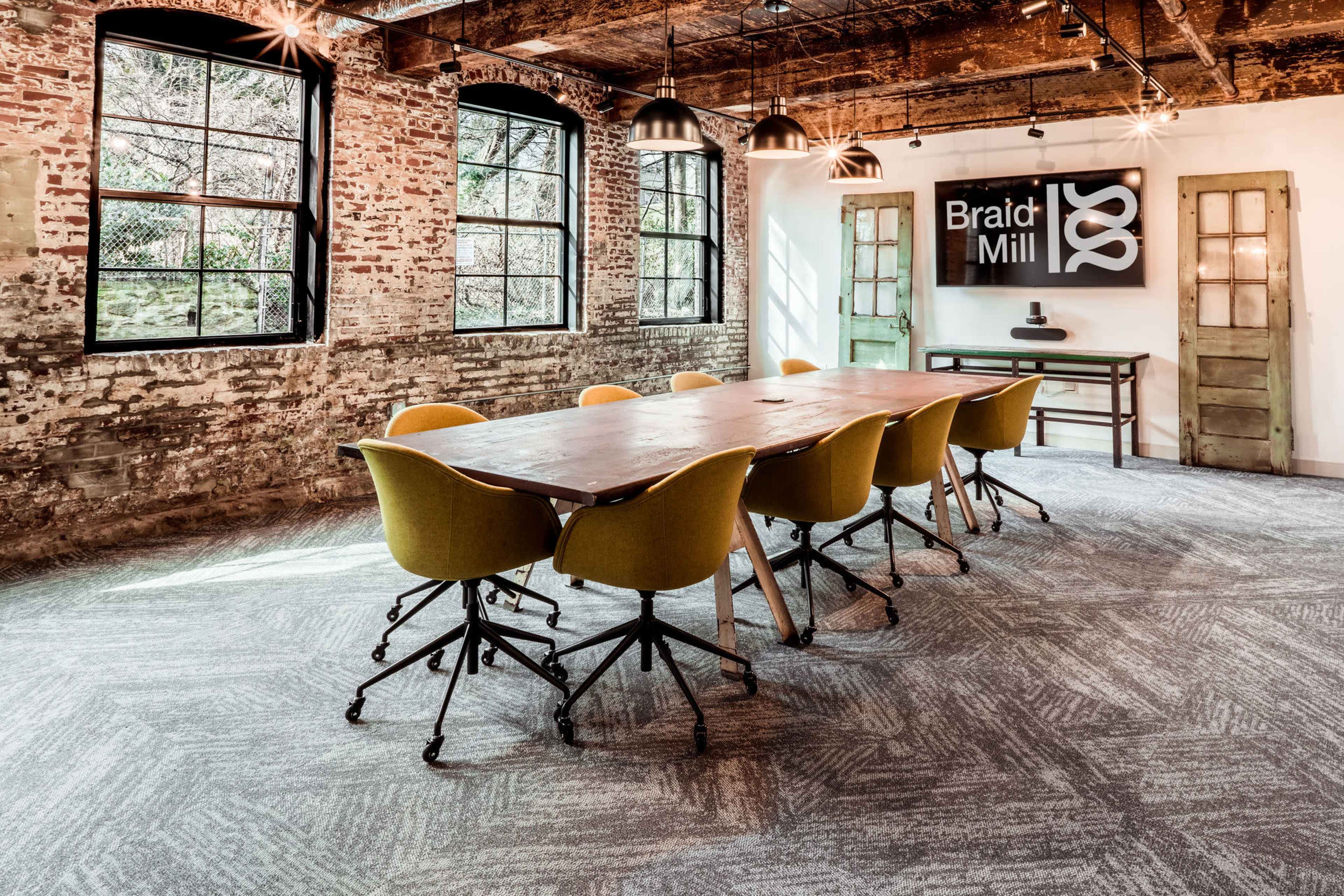 A modern meeting room featuring a large wooden table surrounded by yellow chairs, with exposed brick walls and large windows allowing natural light.