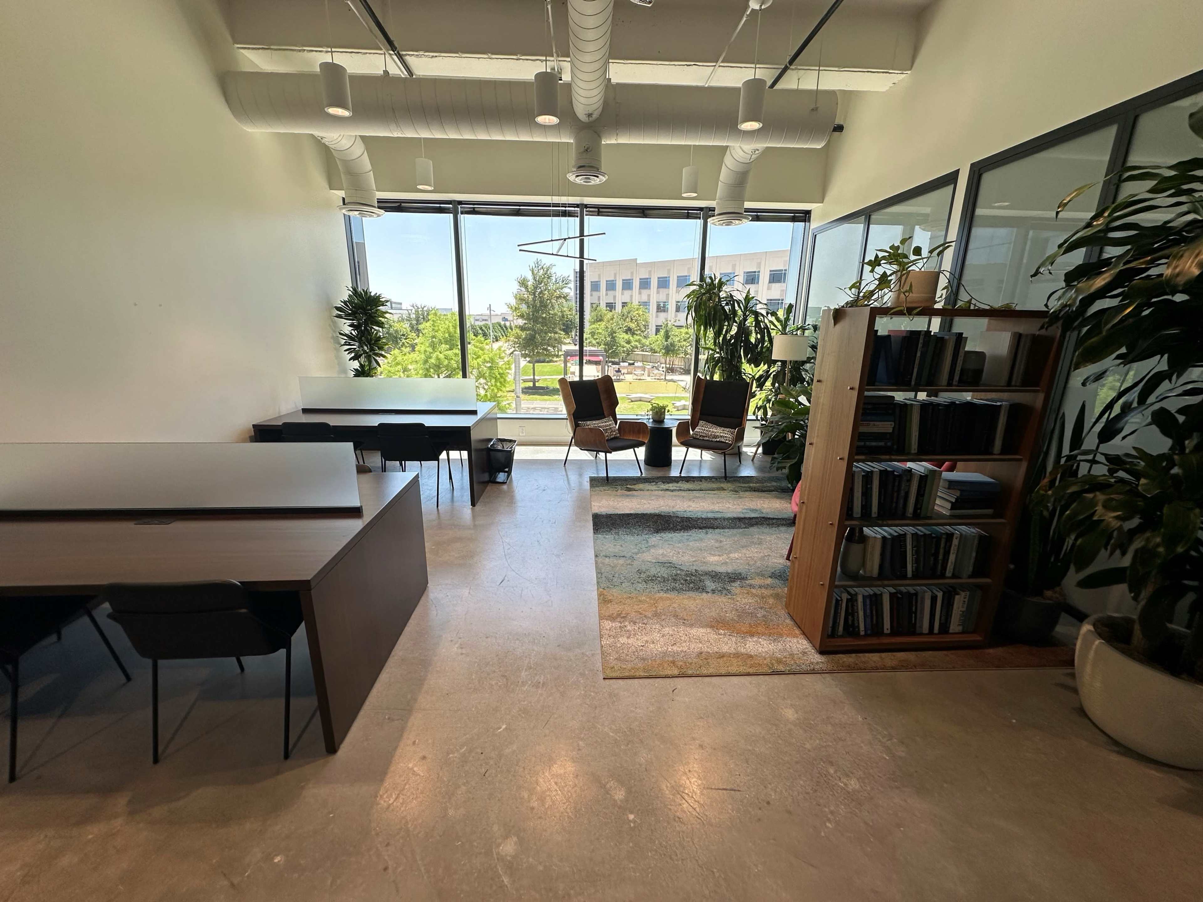 An open office space with a desk area, a bookshelf filled with books, two chairs, and large windows letting in natural light.