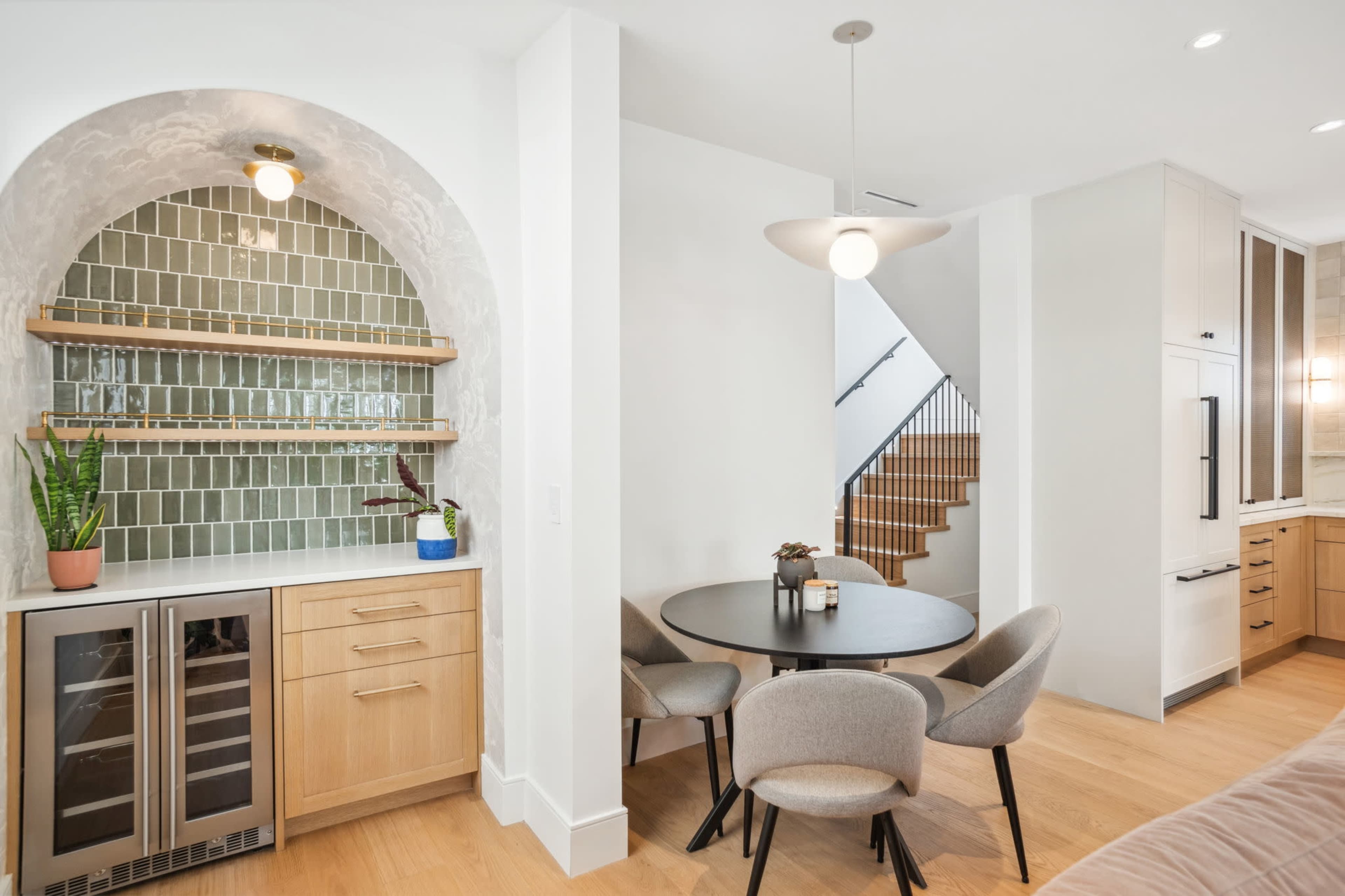 The image shows a modern kitchen area featuring a round dining table with four chairs, a cabinetry section with a wine cooler, and an arched backsplash adorned with green tiles.