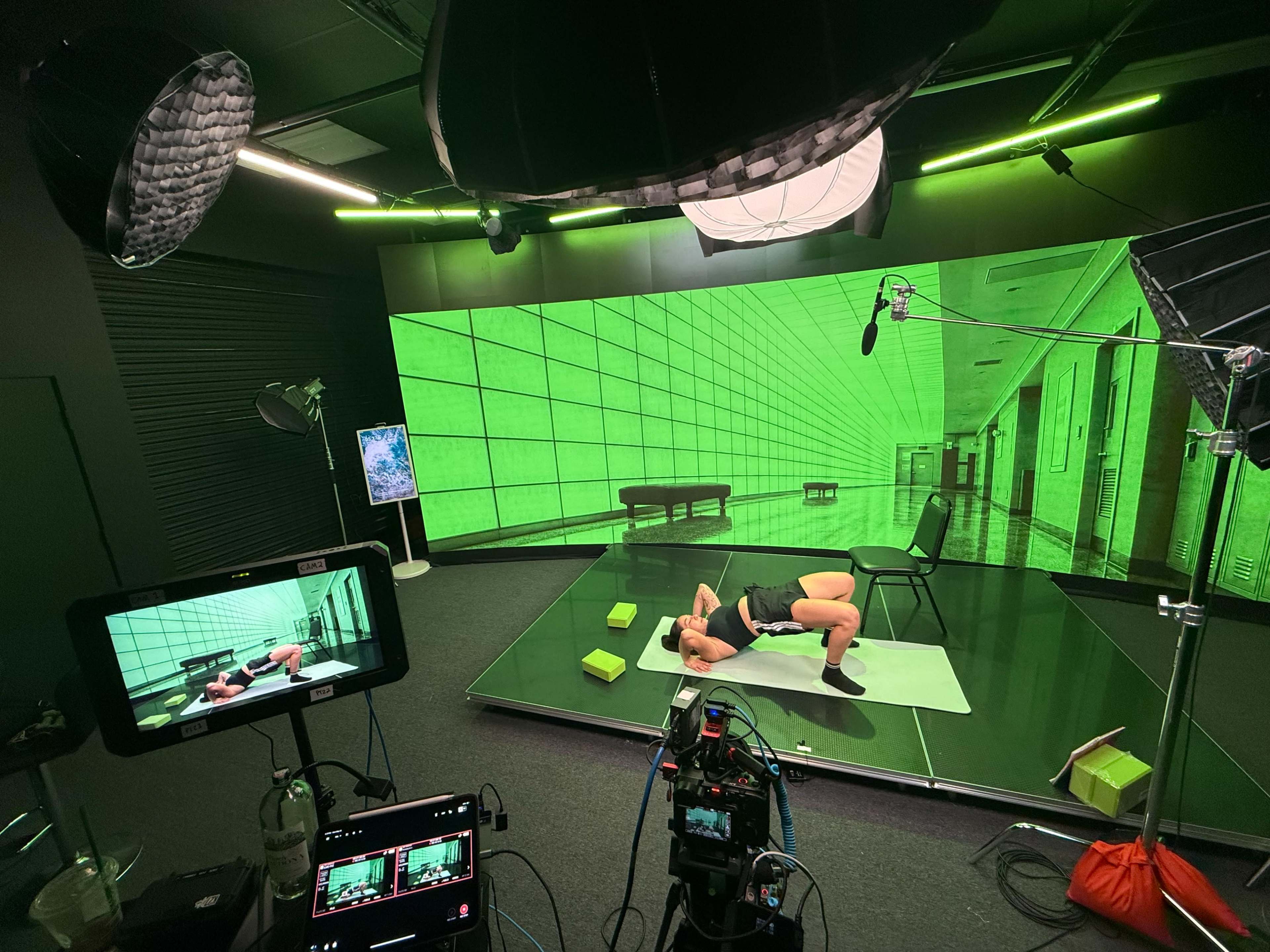A person performs an exercise on a mat in front of a large green screen in a studio setting, with cameras and lighting equipment arranged nearby.