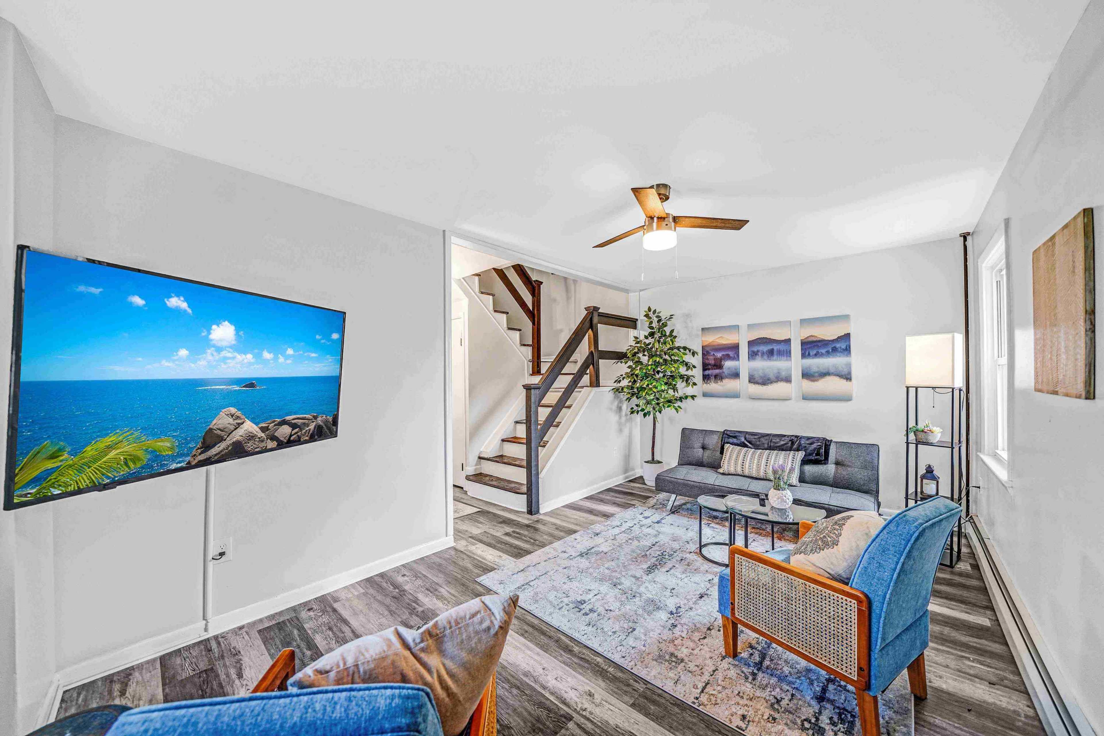 A modern living room with a large television on the wall, a sofa and armchairs, a staircase in the background, and decorative artwork.