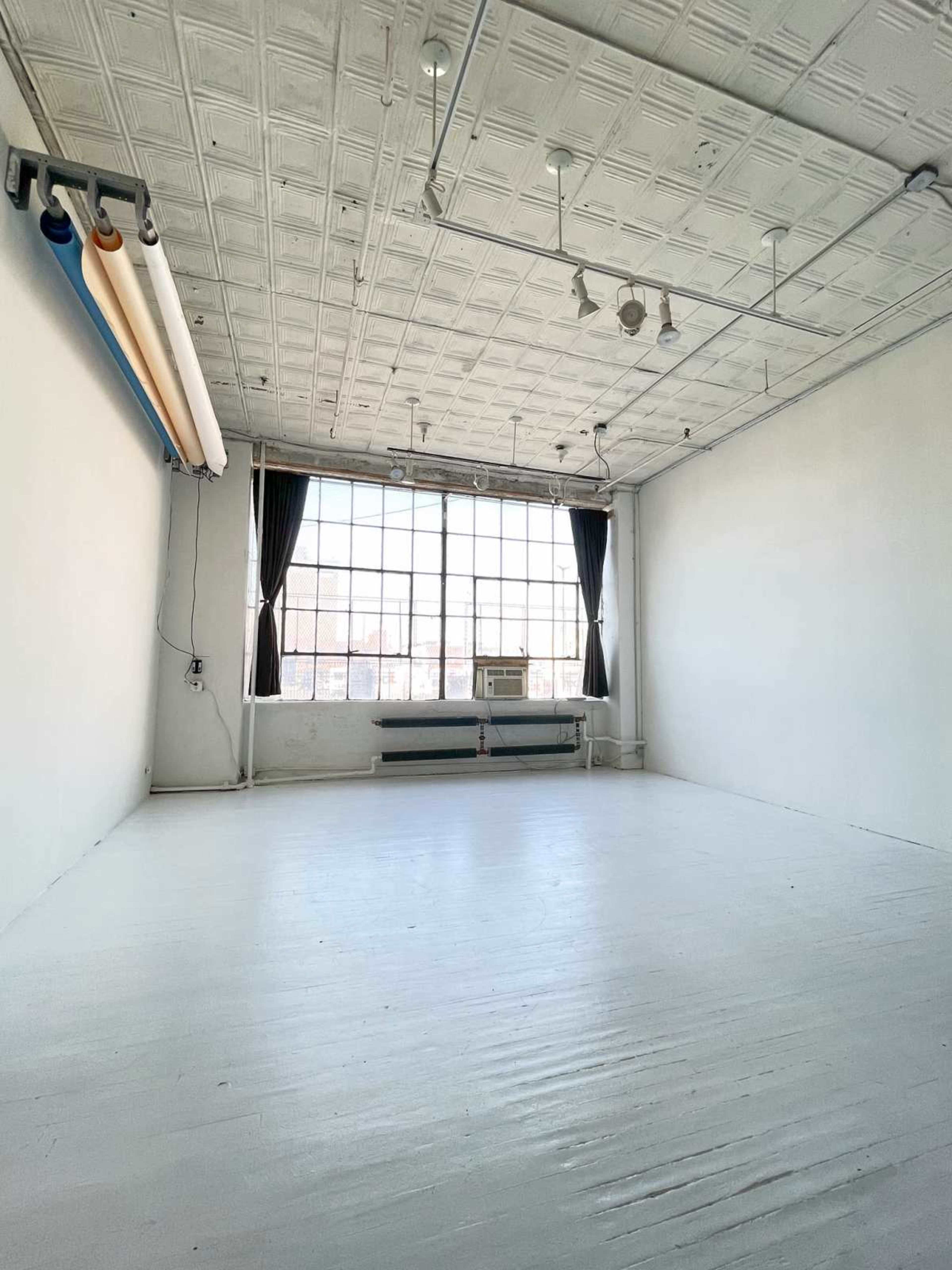 The image shows an empty, well-lit studio space with large windows, white walls, and a simple ceiling design.