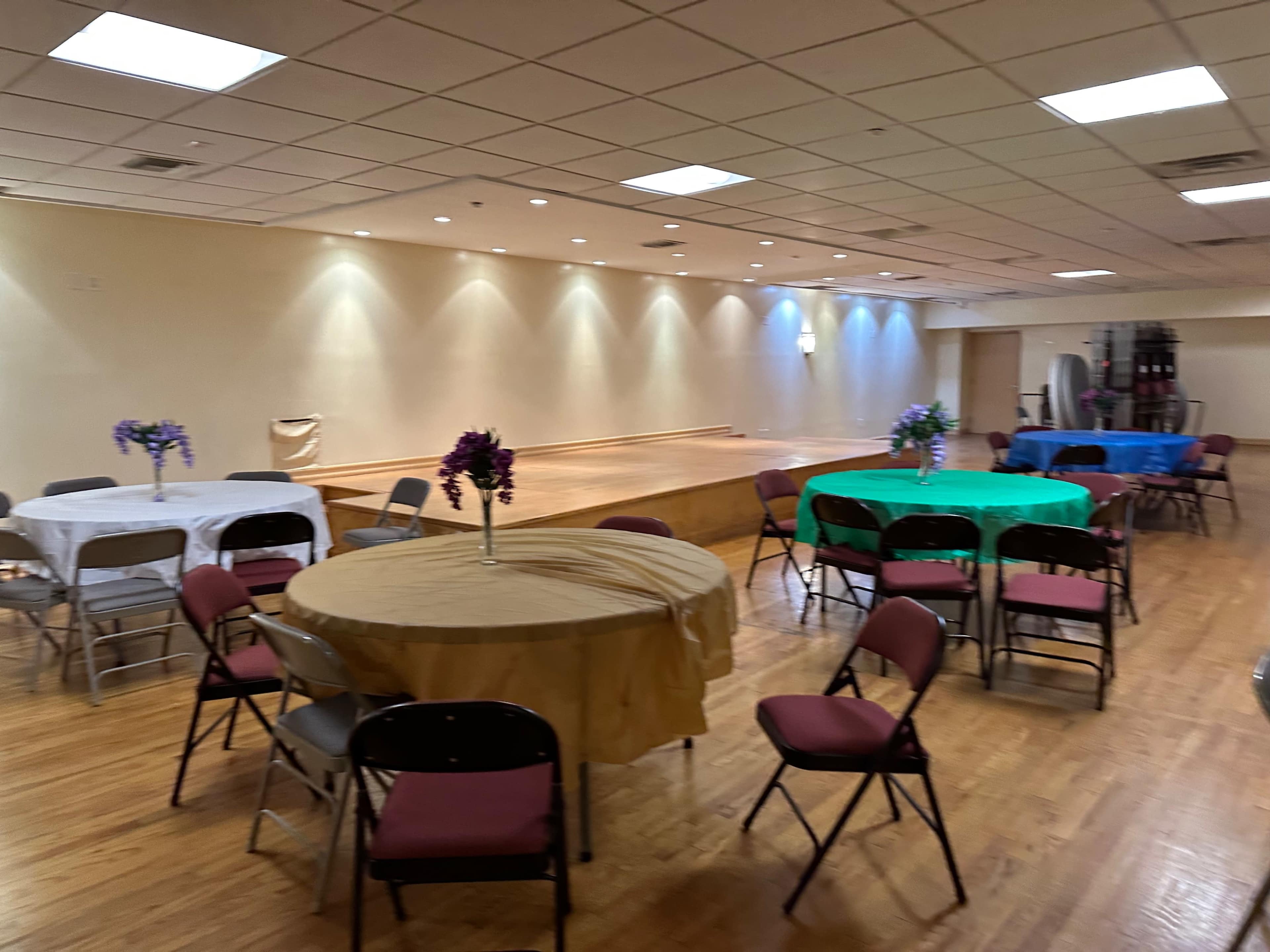 A spacious event room features several round tables with different colored tablecloths and chairs arranged around them.