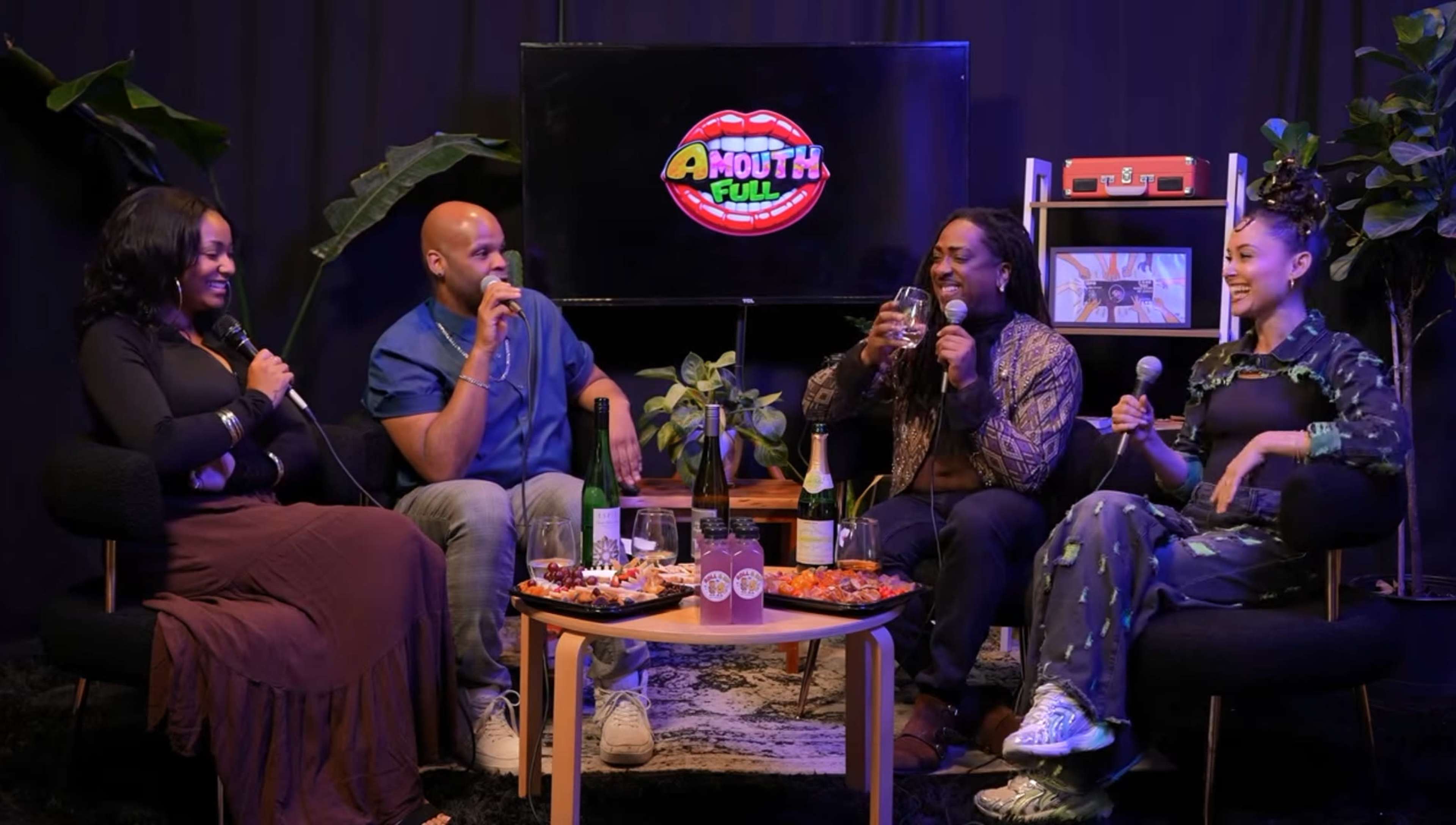 A group of four people sit around a coffee table filled with food and drinks, engaging in conversation while microphones are positioned in front of them.