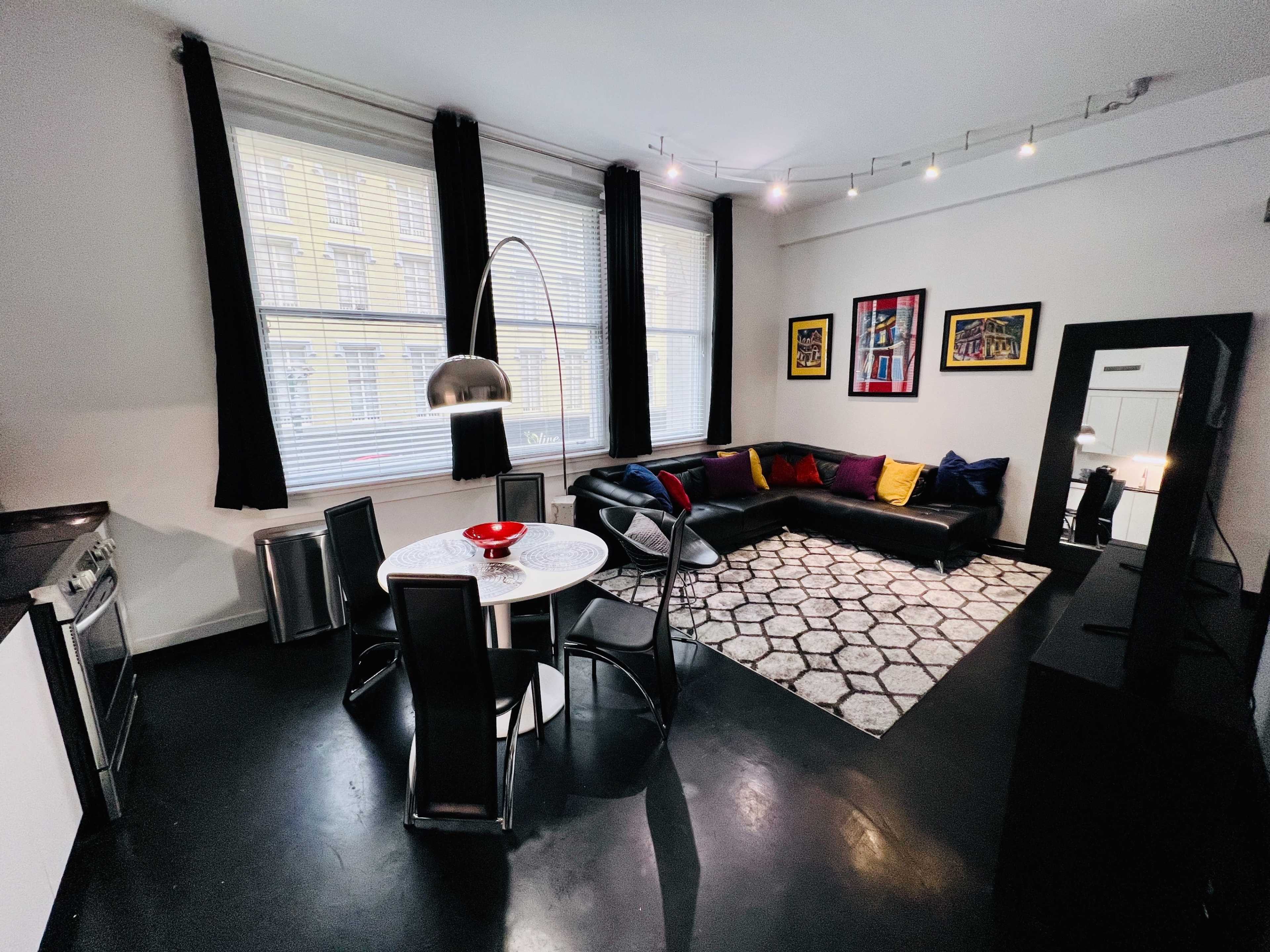 The image shows a modern living space featuring a black sectional sofa, a round dining table with chairs, and large windows with white blinds.