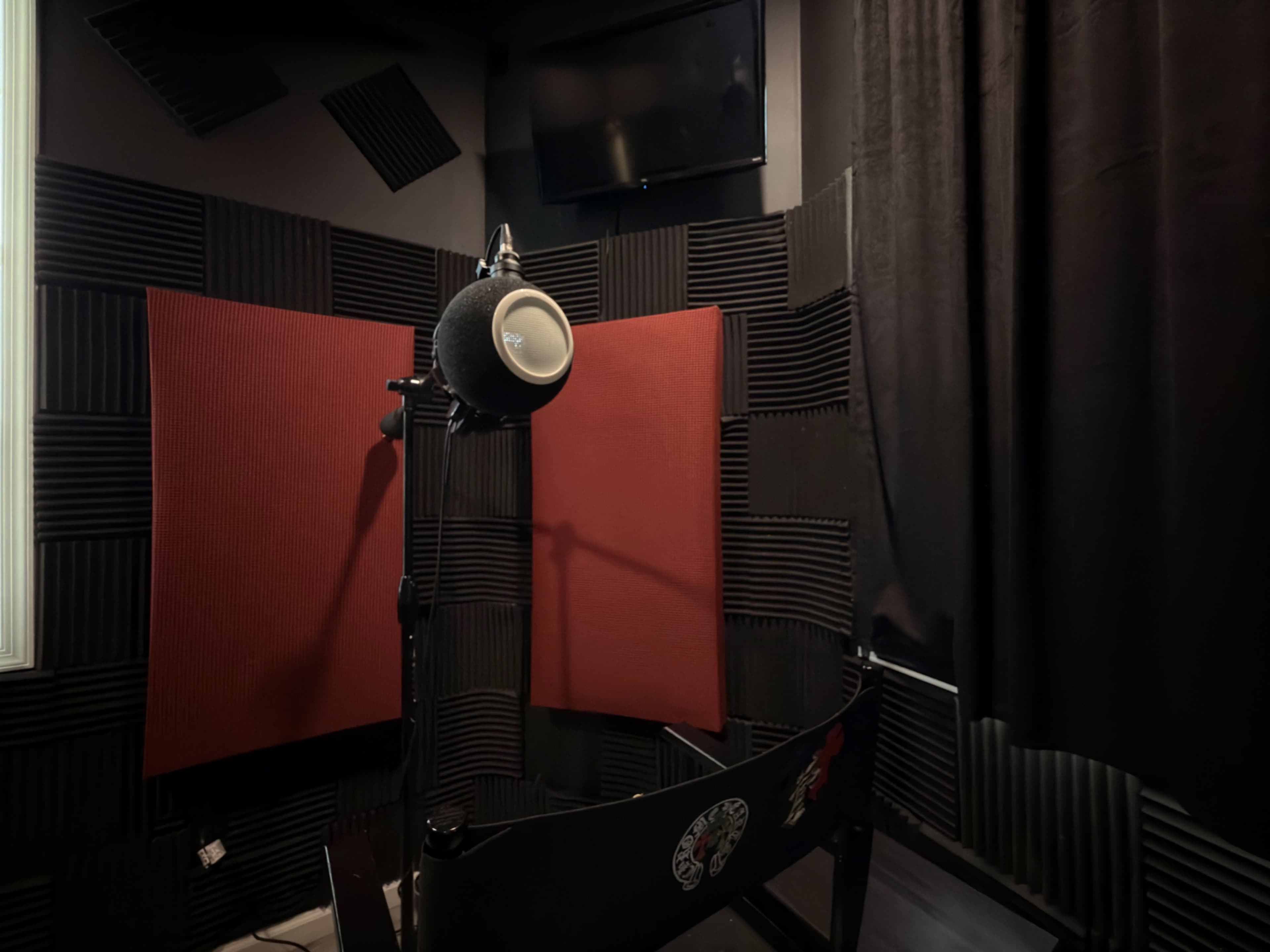 The image shows a soundproof recording booth with a microphone mounted on a stand, surrounded by red acoustic panels and dark curtains.