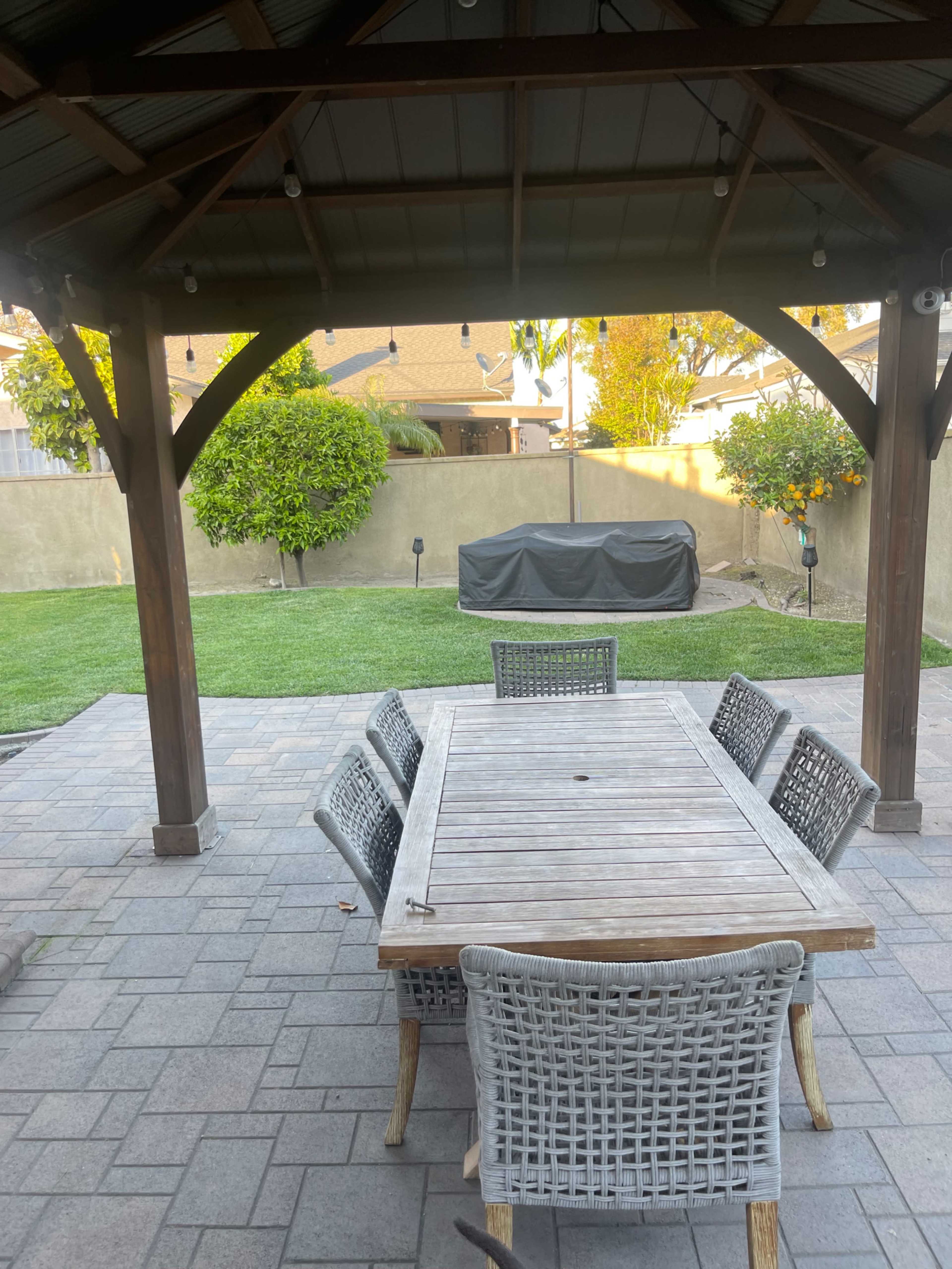 A wooden dining table with six wicker chairs is placed under a wooden gazebo in a backyard featuring grass and a BBQ grill covered in a tarp.