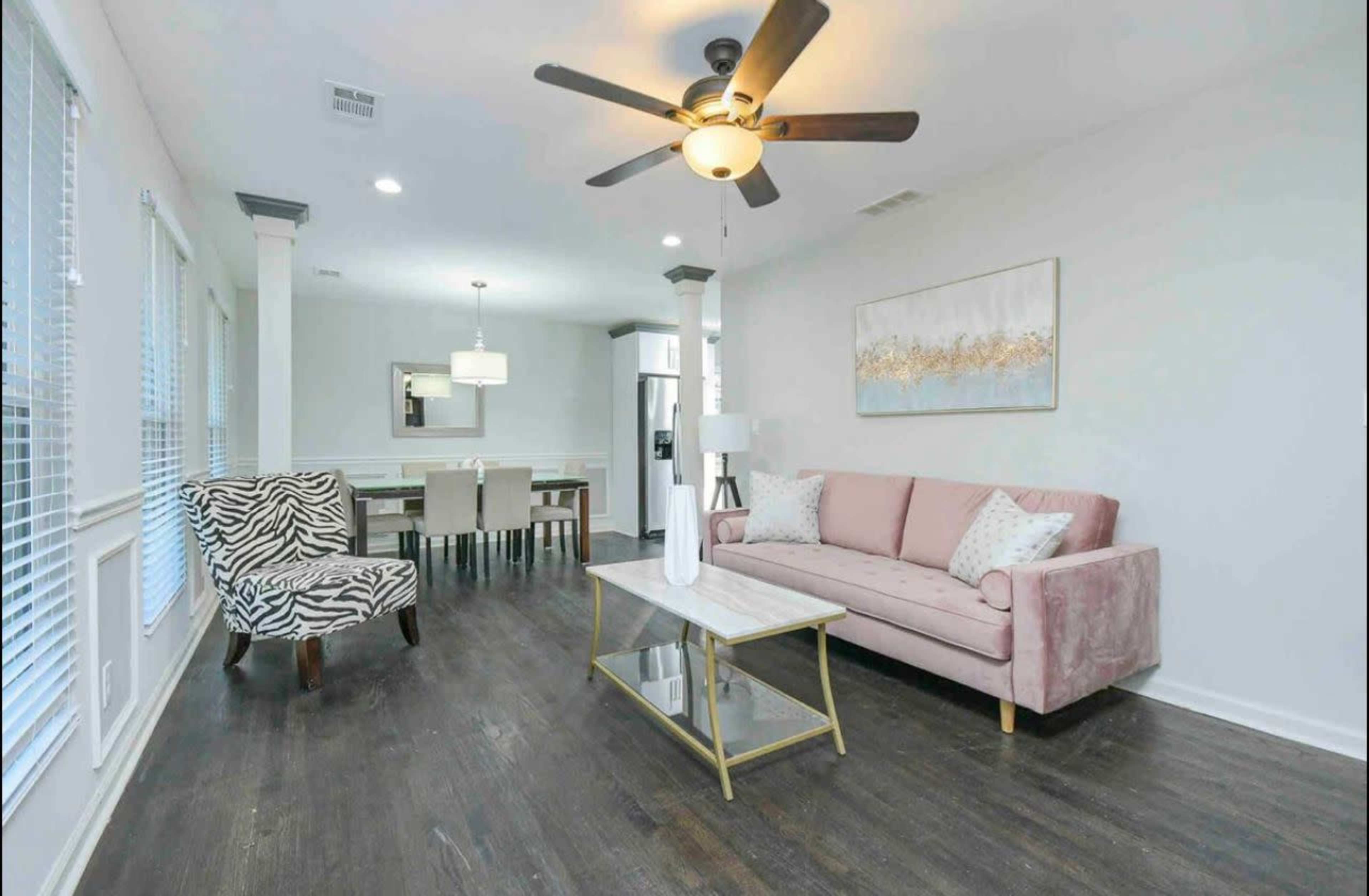 The image shows a modern living room with a pink sofa, a zebra-print armchair, a coffee table, and a dining area in the background.