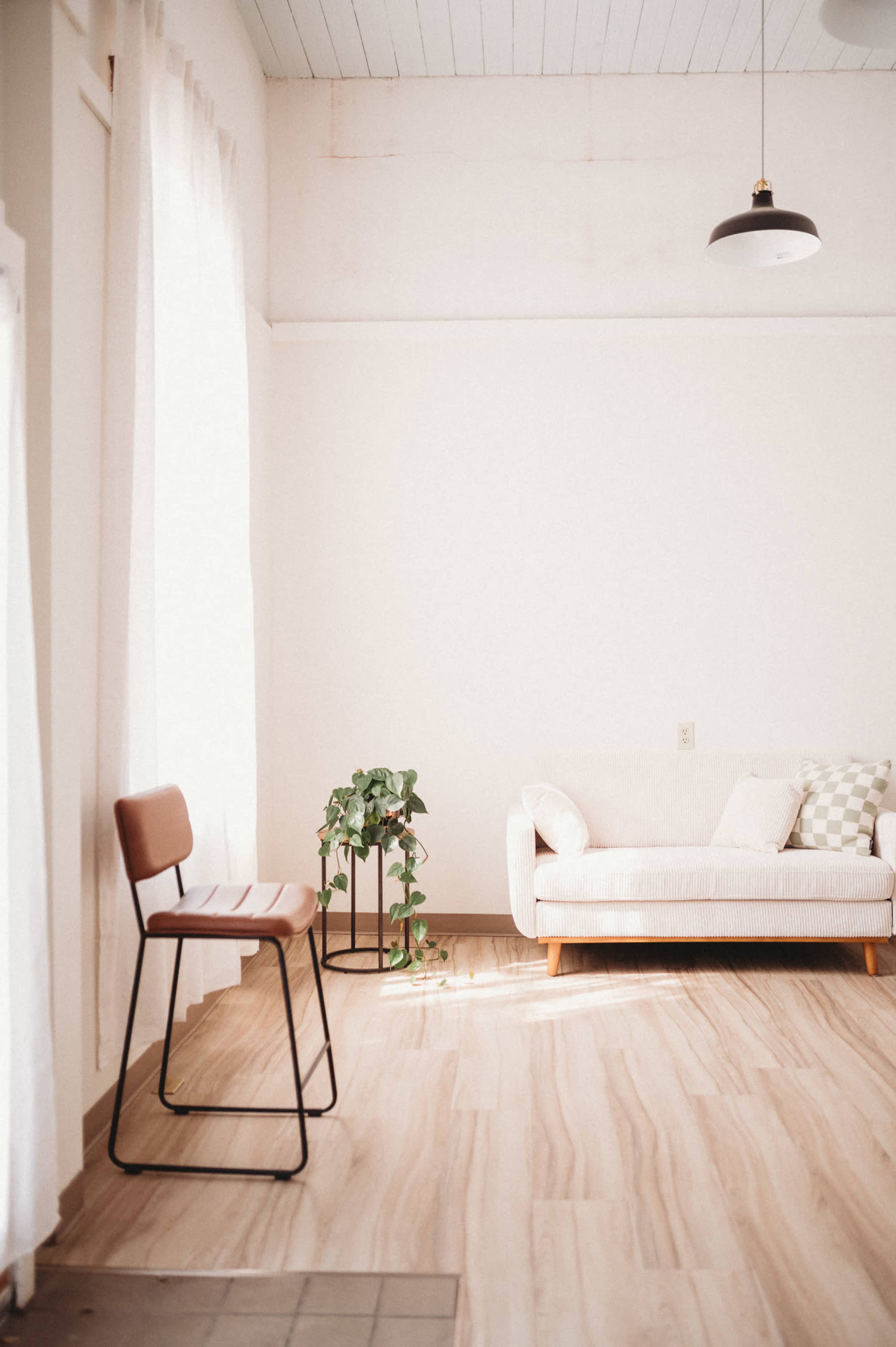 The image shows a minimalistic living room featuring a light-colored couch, a small potted plant, and a simple chair against a wall with large windows.