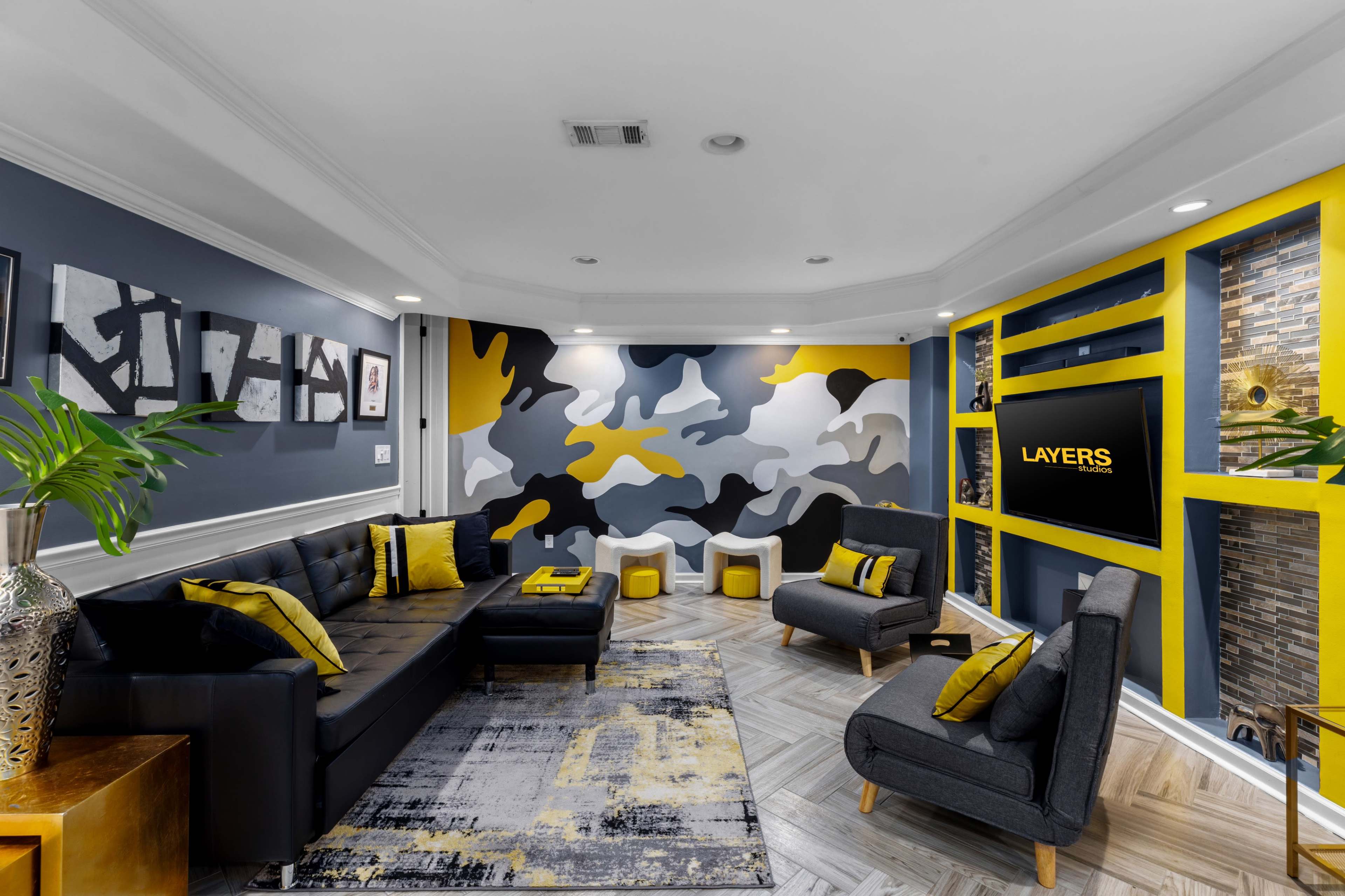 A modern living room features a black sofa, yellow accents, abstract wall art, and a camouflage-patterned feature wall with a TV.
