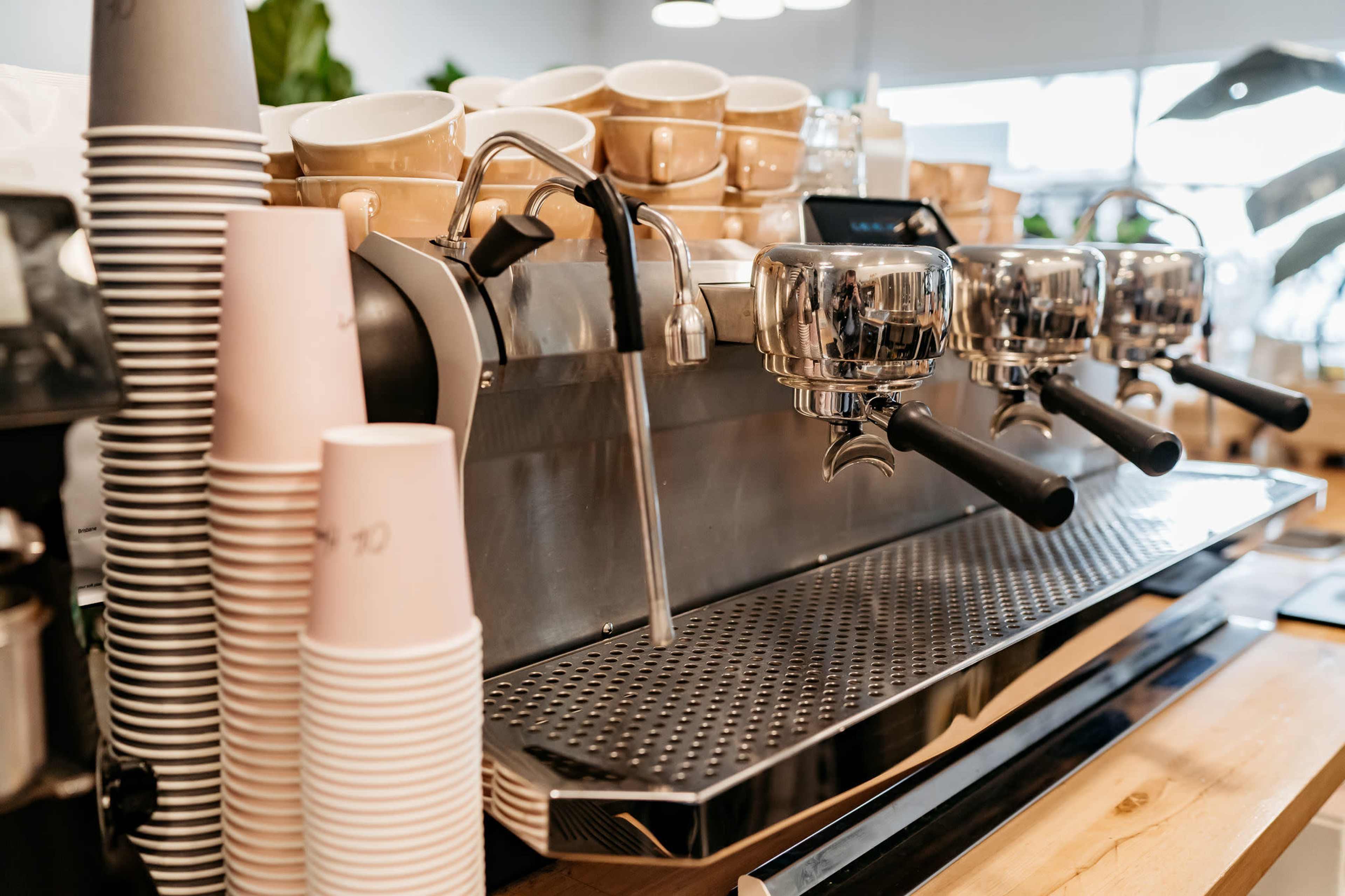 A coffee machine with three group heads sits on a countertop next to stacks of cups and disposable coffee cups.