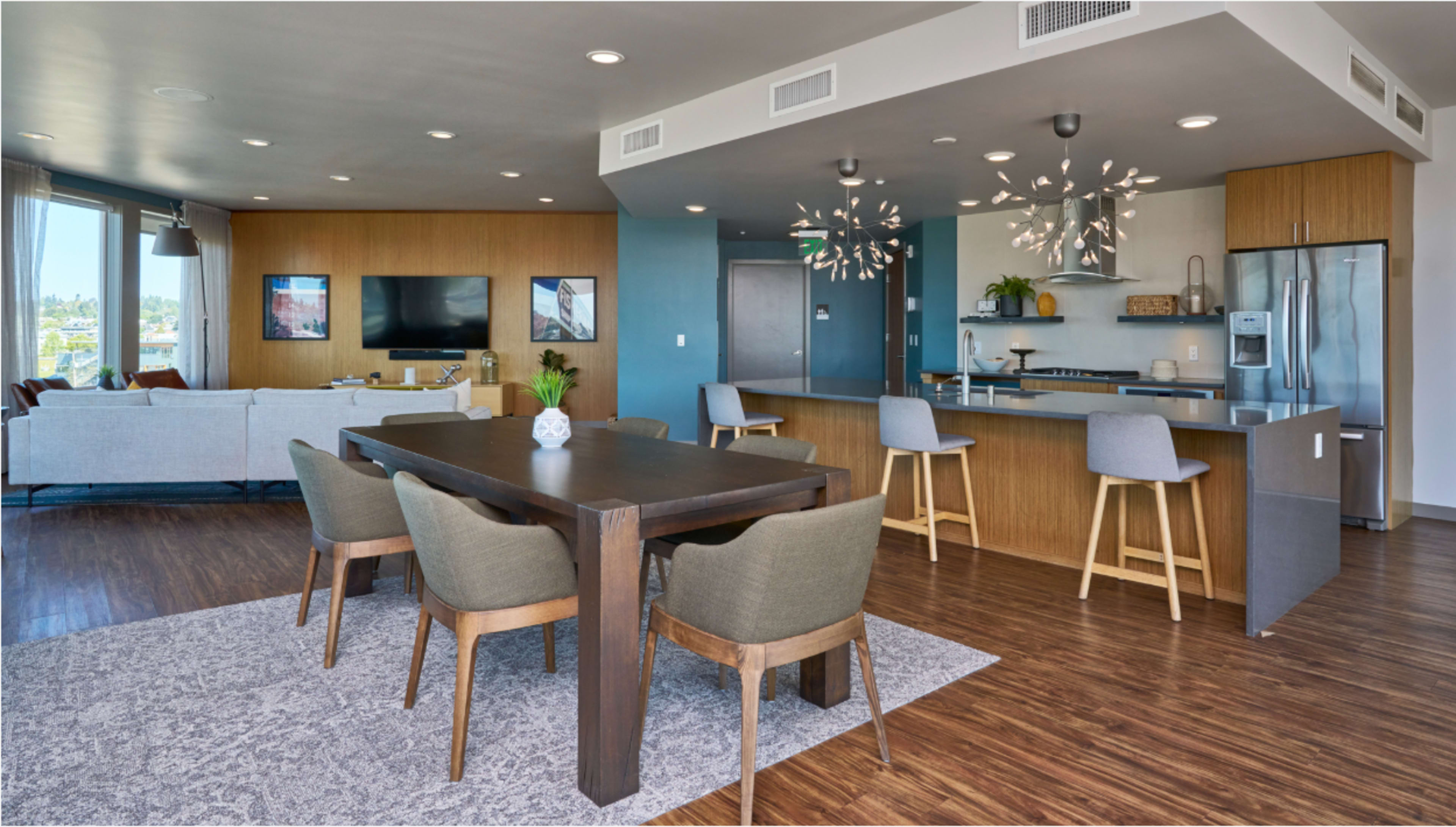The image shows a modern open-concept living space featuring a dining area with a wooden table and upholstered chairs, adjacent to a kitchen with stainless steel appliances and a living room with a sofa and television.
