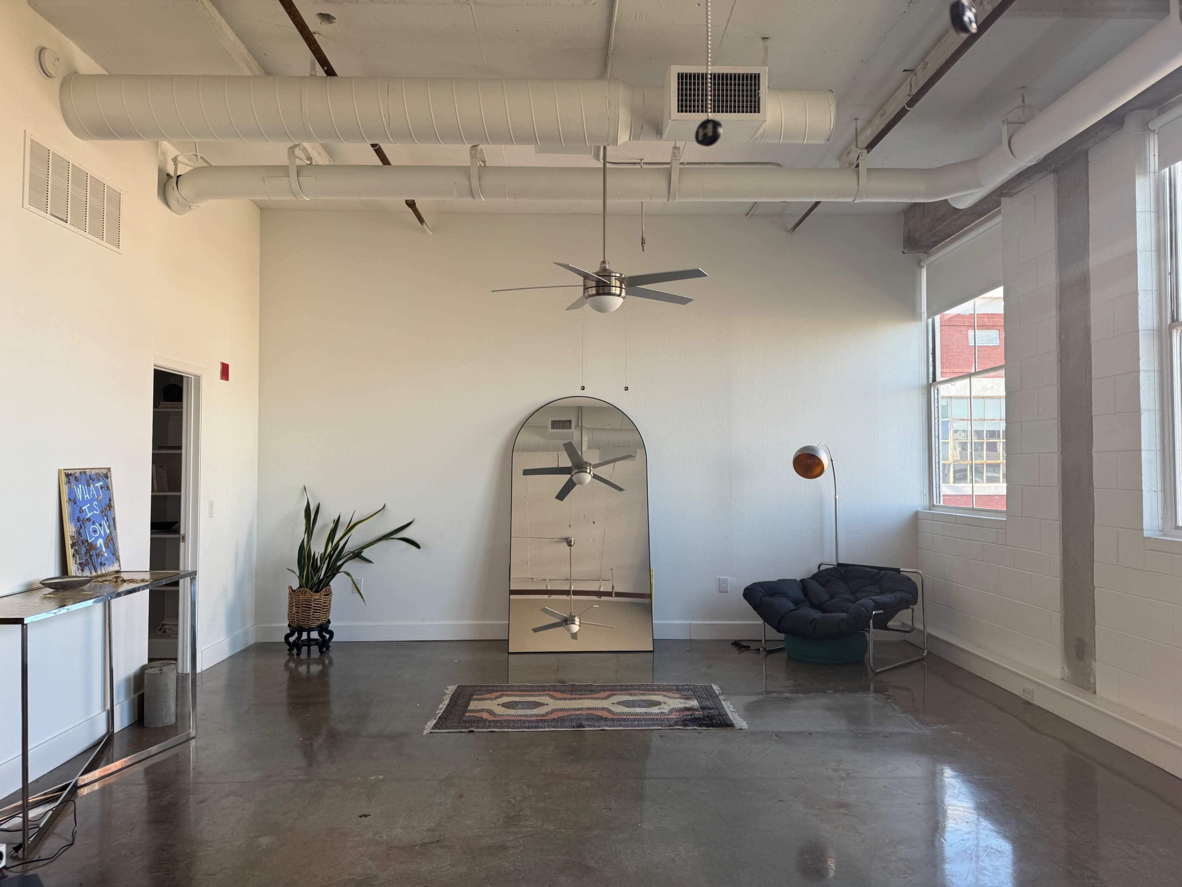 The image shows a spacious, modern room with concrete floors, a large mirror, a ceiling fan, a potted plant, and a black lounge chair beside a floor lamp.