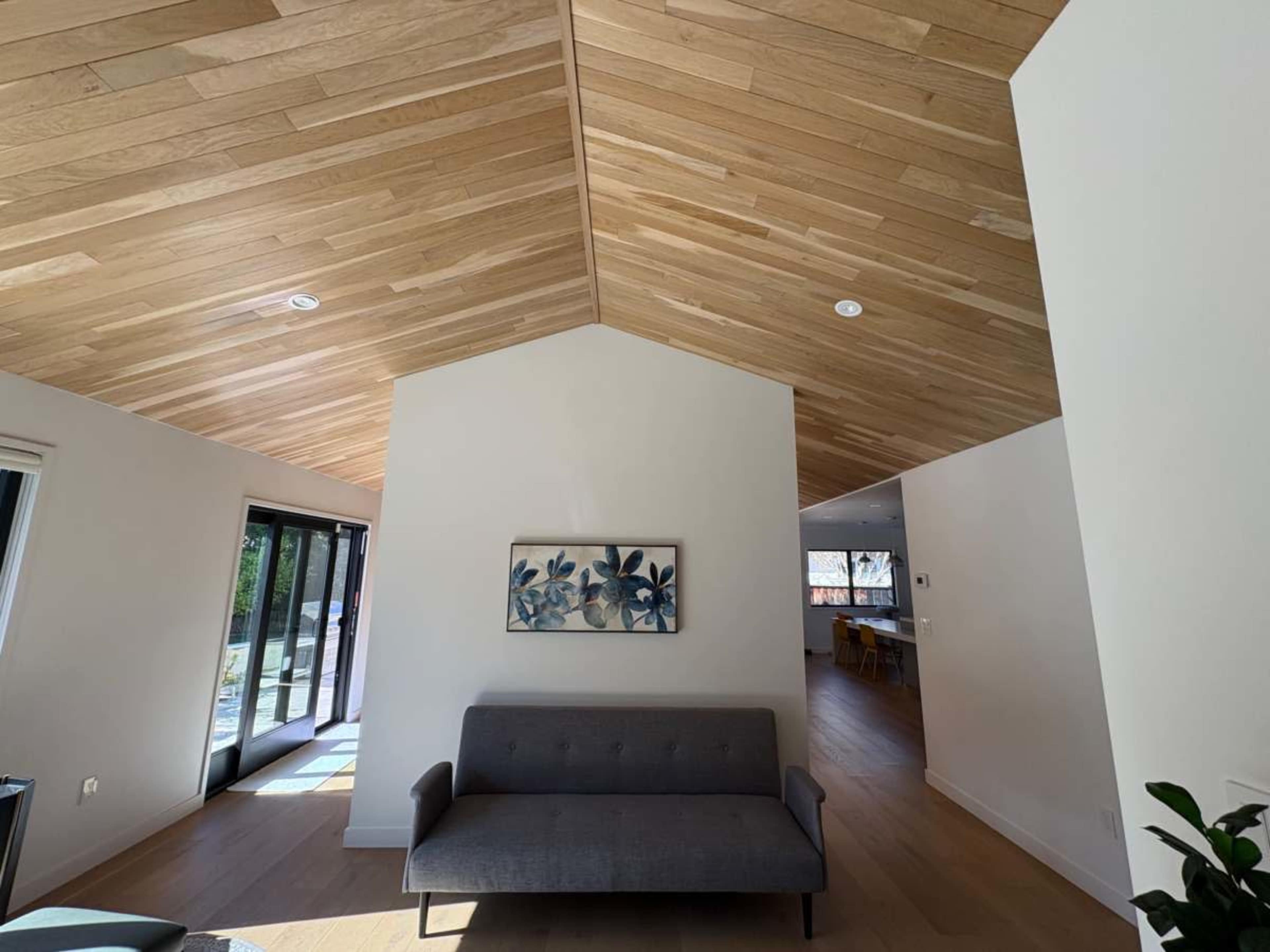 A modern interior space features a wooden ceiling with a peak, a gray sofa against a white wall, and sliding doors leading outside.