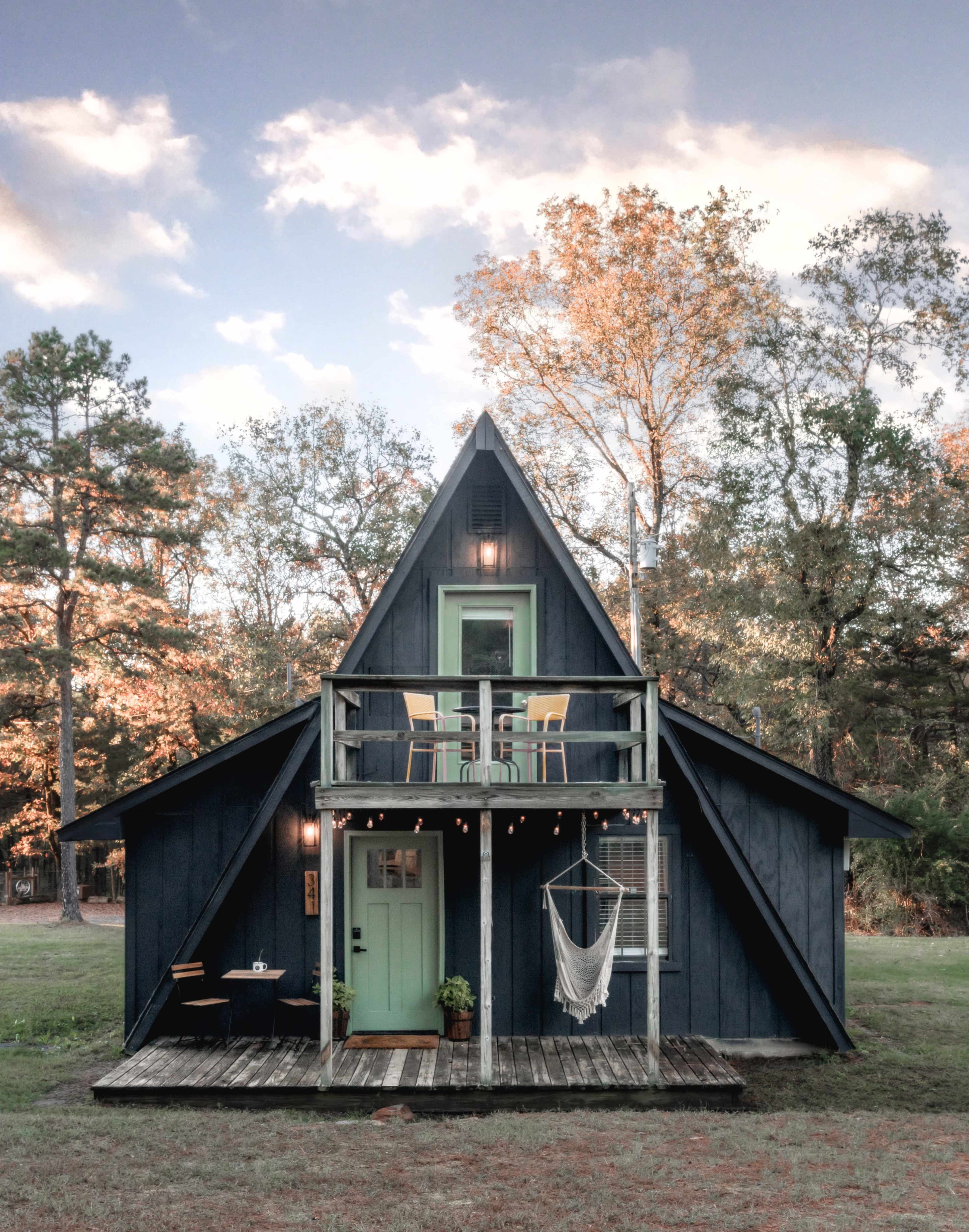 A triangular-roofed cabin with a green door and balcony is surrounded by trees and has outdoor seating.