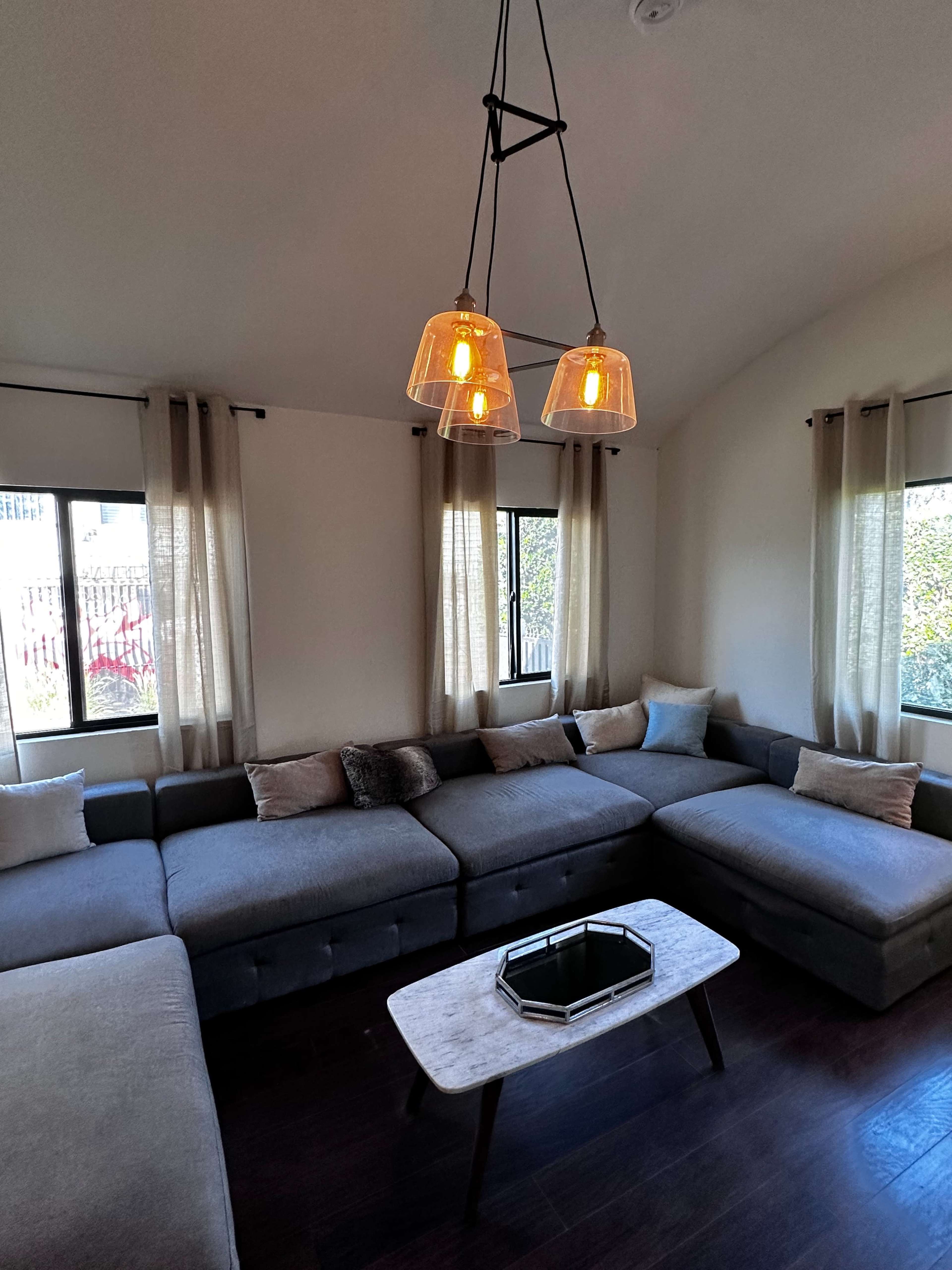 The image shows a modern living room with a large sectional sofa, a marble coffee table, and pendant lighting, complemented by large windows dressed with curtains.
