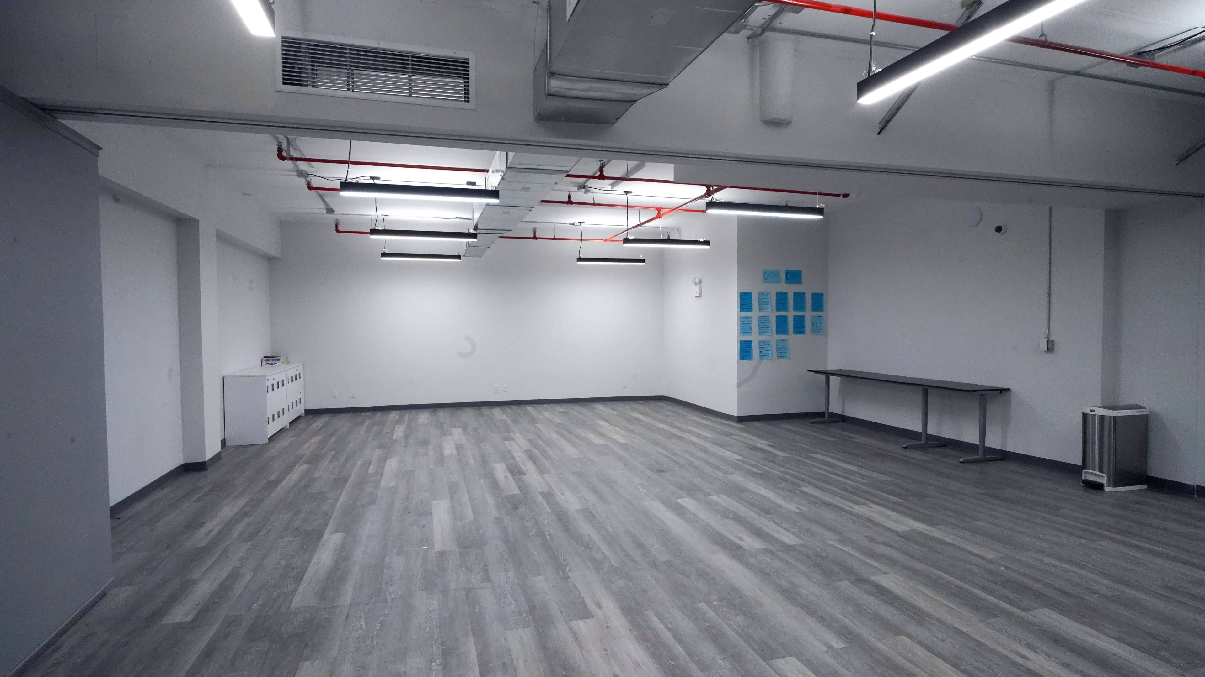 The image shows an empty room with gray walls, wooden flooring, and overhead fluorescent lighting.