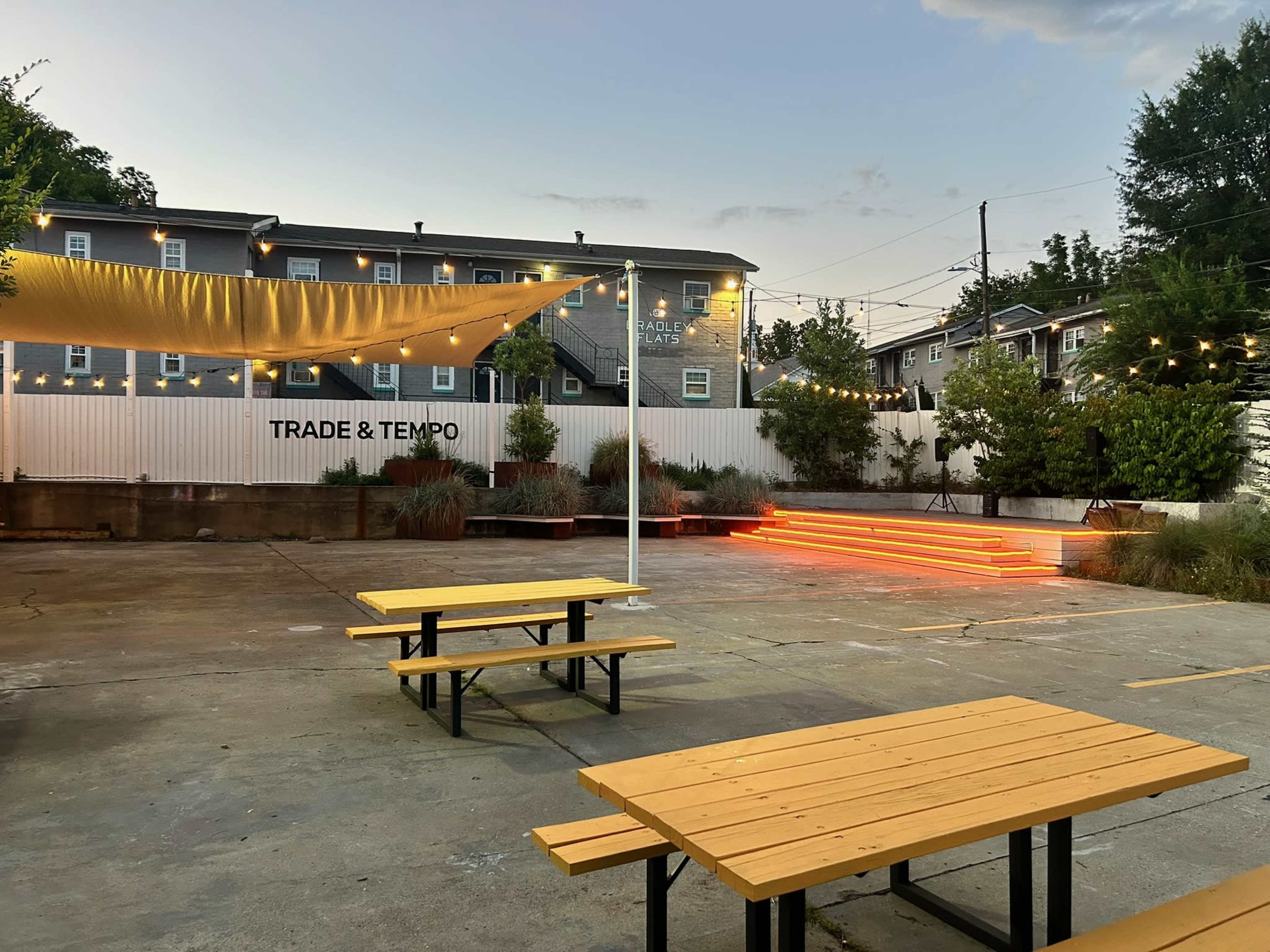 The image shows an outdoor area with several wooden picnic tables, an illuminated stage, and a white backdrop featuring the text "TRADE & TEMPO," surrounded by trees and string lights.