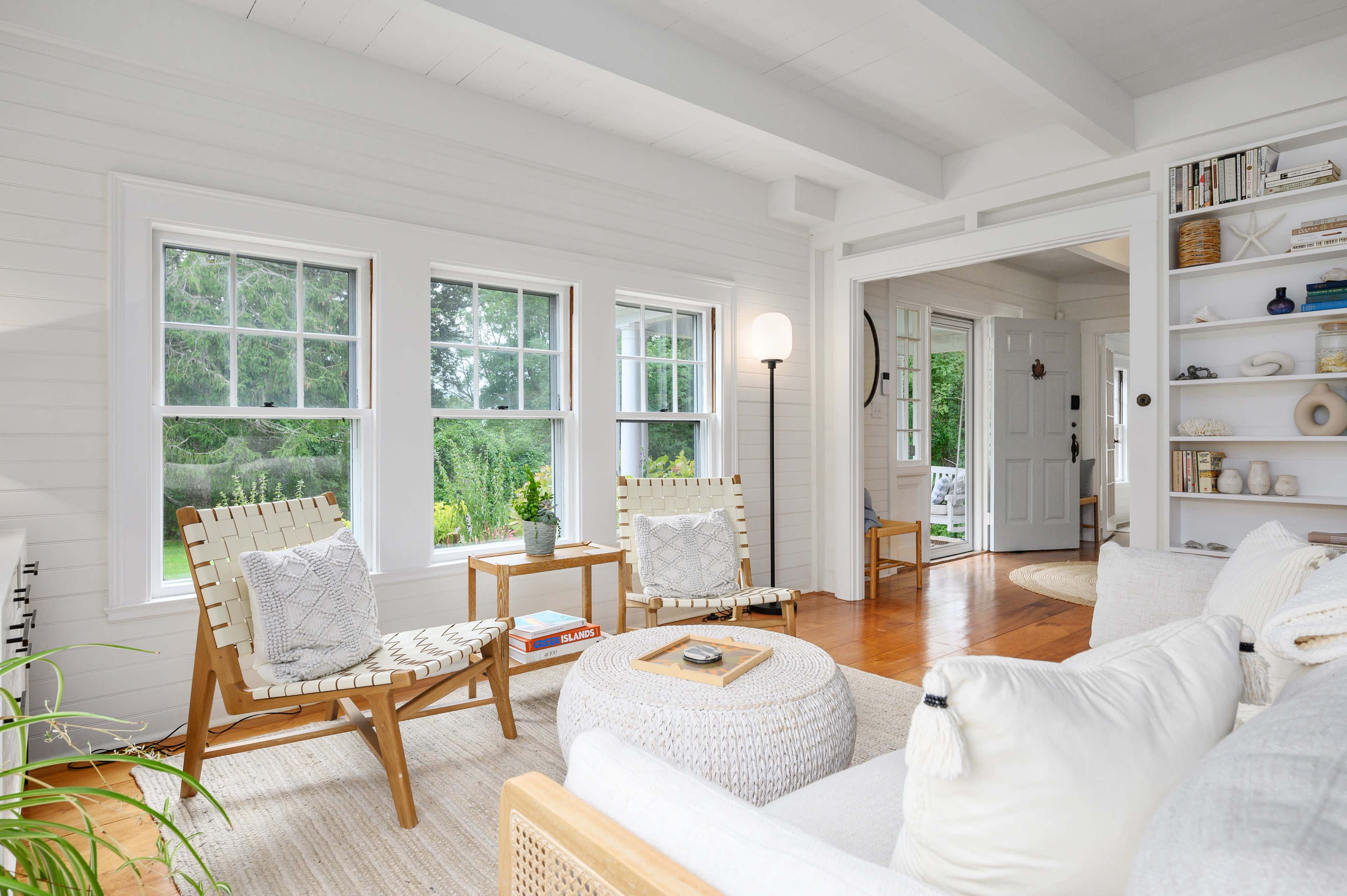A bright and airy living room with large windows, two wooden chairs, a round ottoman, and a cozy sofa.