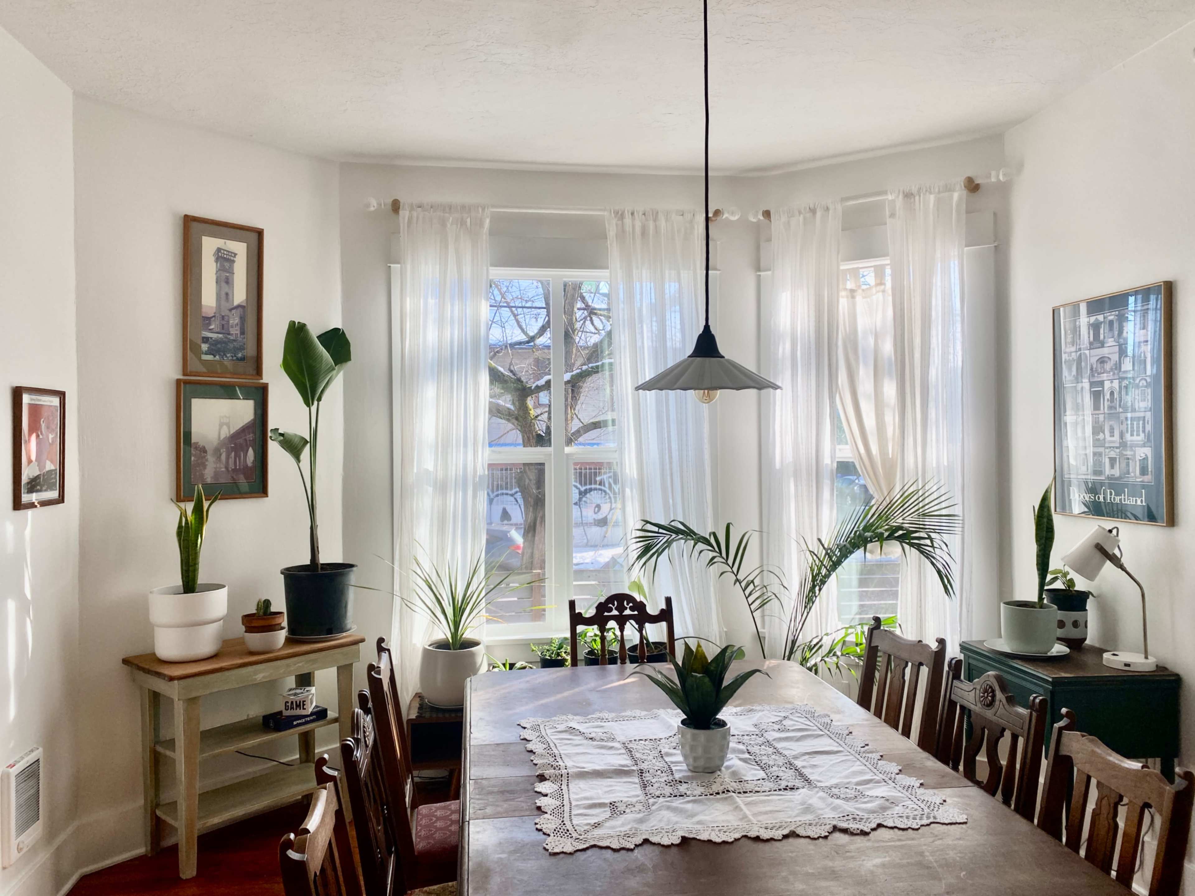 A bright dining room features a large wooden table surrounded by chairs, with plants and natural light coming through the windows.