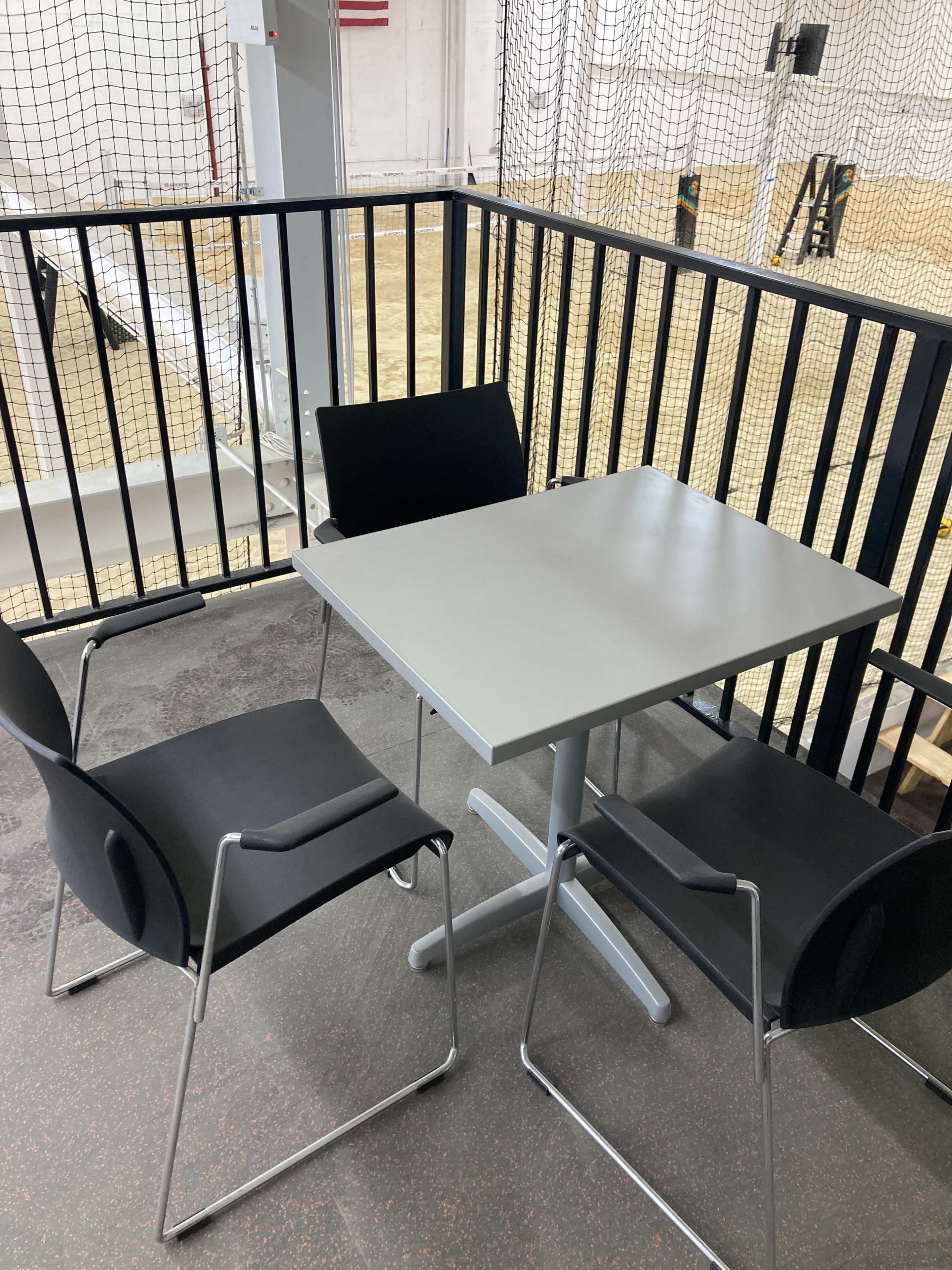 A gray table with three black chairs is situated on a balcony overlooking a gymnasium court.