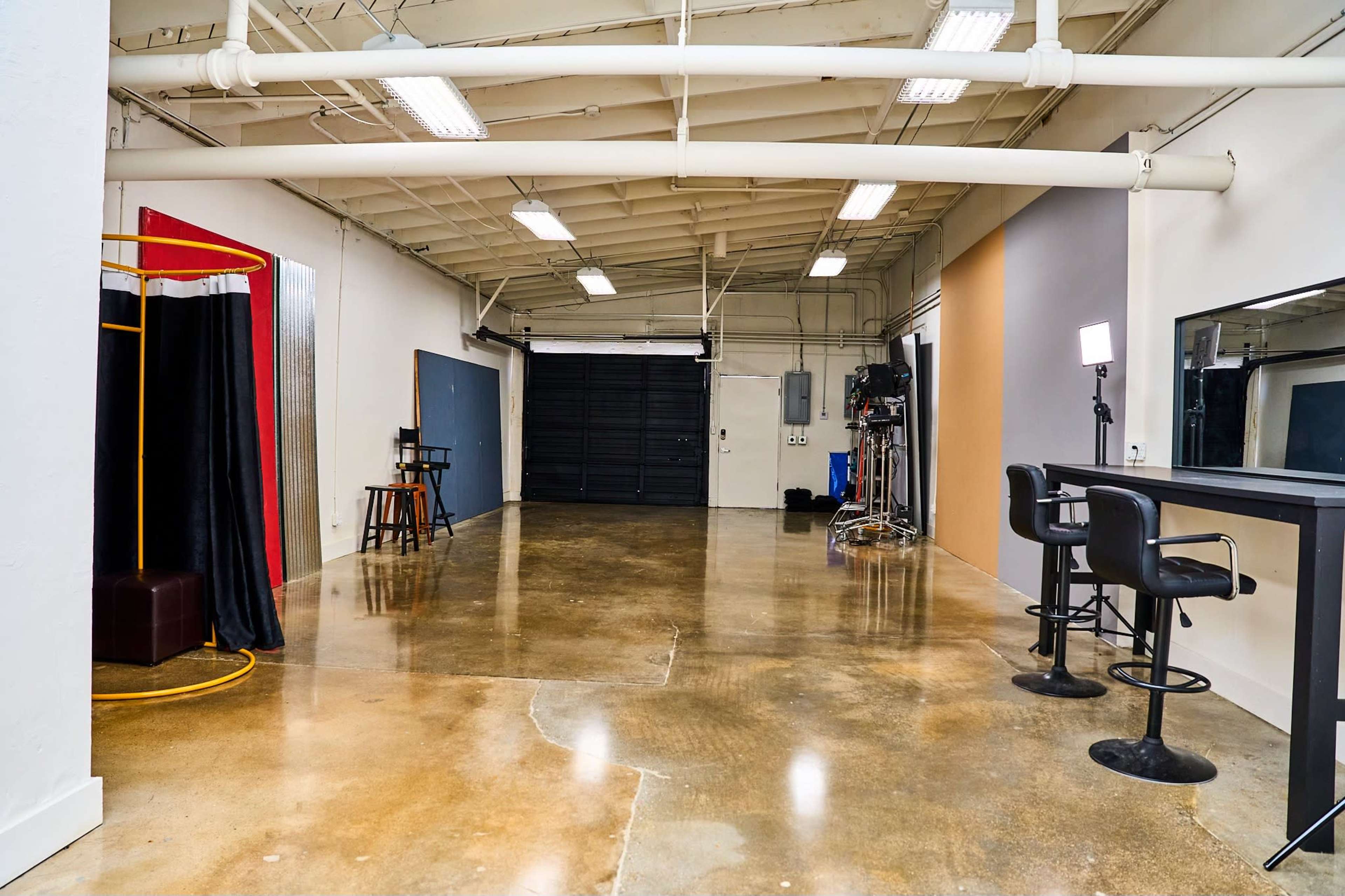 An empty interior space with polished concrete floors, featuring a black roll-up garage door, a mirrored table with chairs, and a photography backdrop setup in one corner.