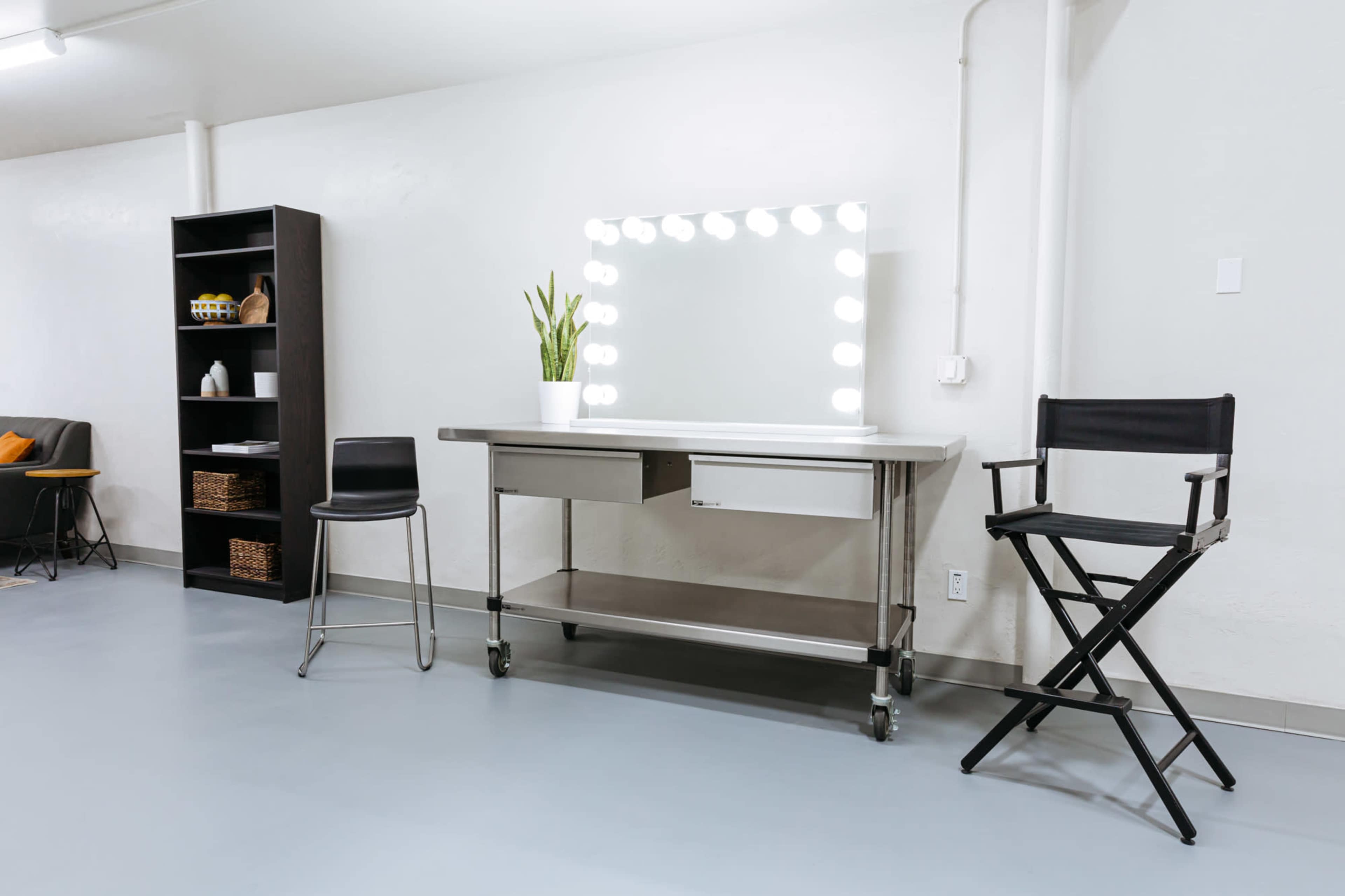 A minimalist makeup vanity with a large illuminated mirror, a black director's chair, and a tall shelving unit in a well-lit, neutral-colored room.
