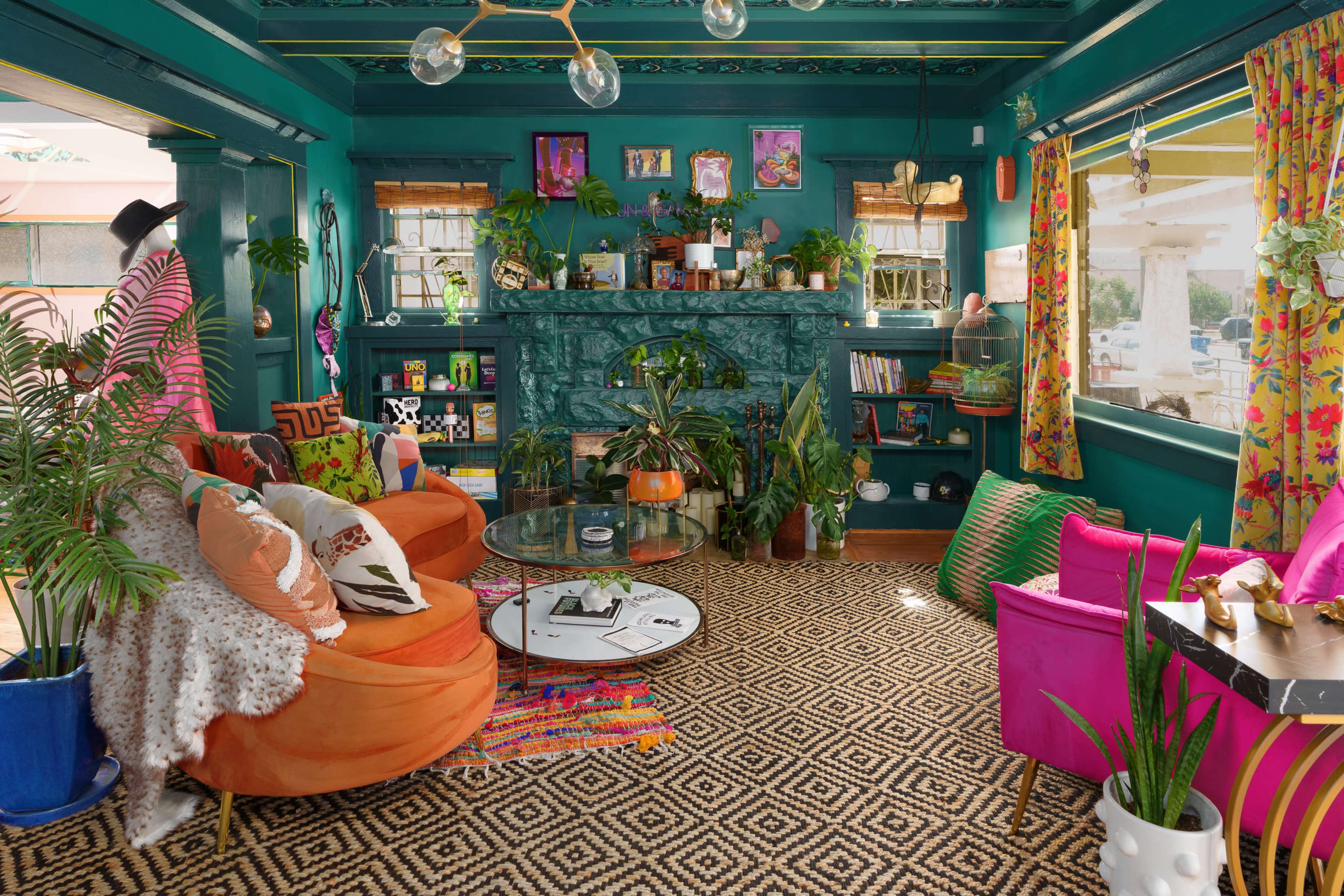 The image shows a vibrant living room with teal walls, a stone fireplace, colorful furniture, and various plants arranged around a circular glass coffee table.