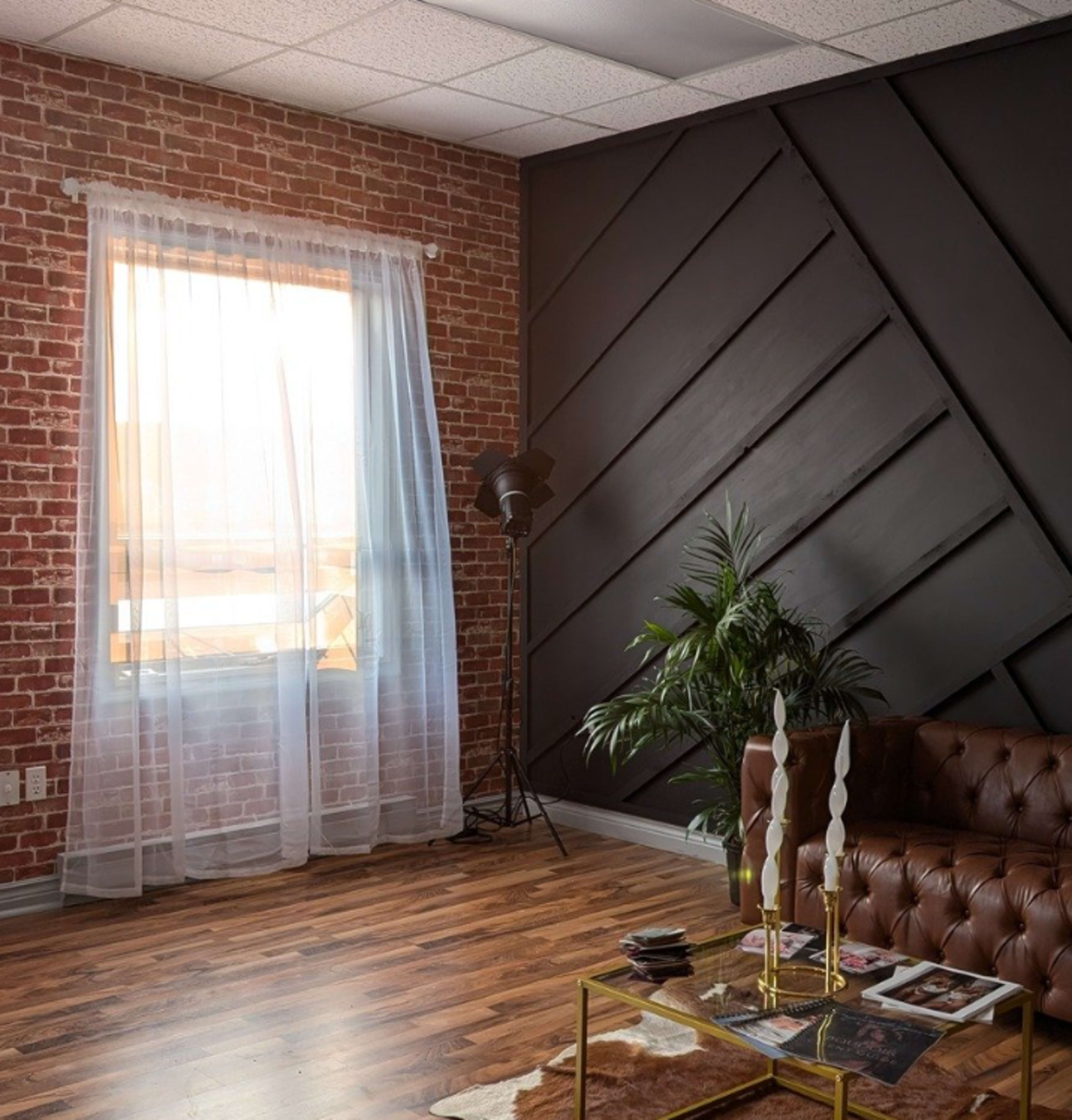 The image shows a stylish interior with a brick wall, a large window draped with sheer curtains, a brown leather sofa, and a tropical plant beside a coffee table.