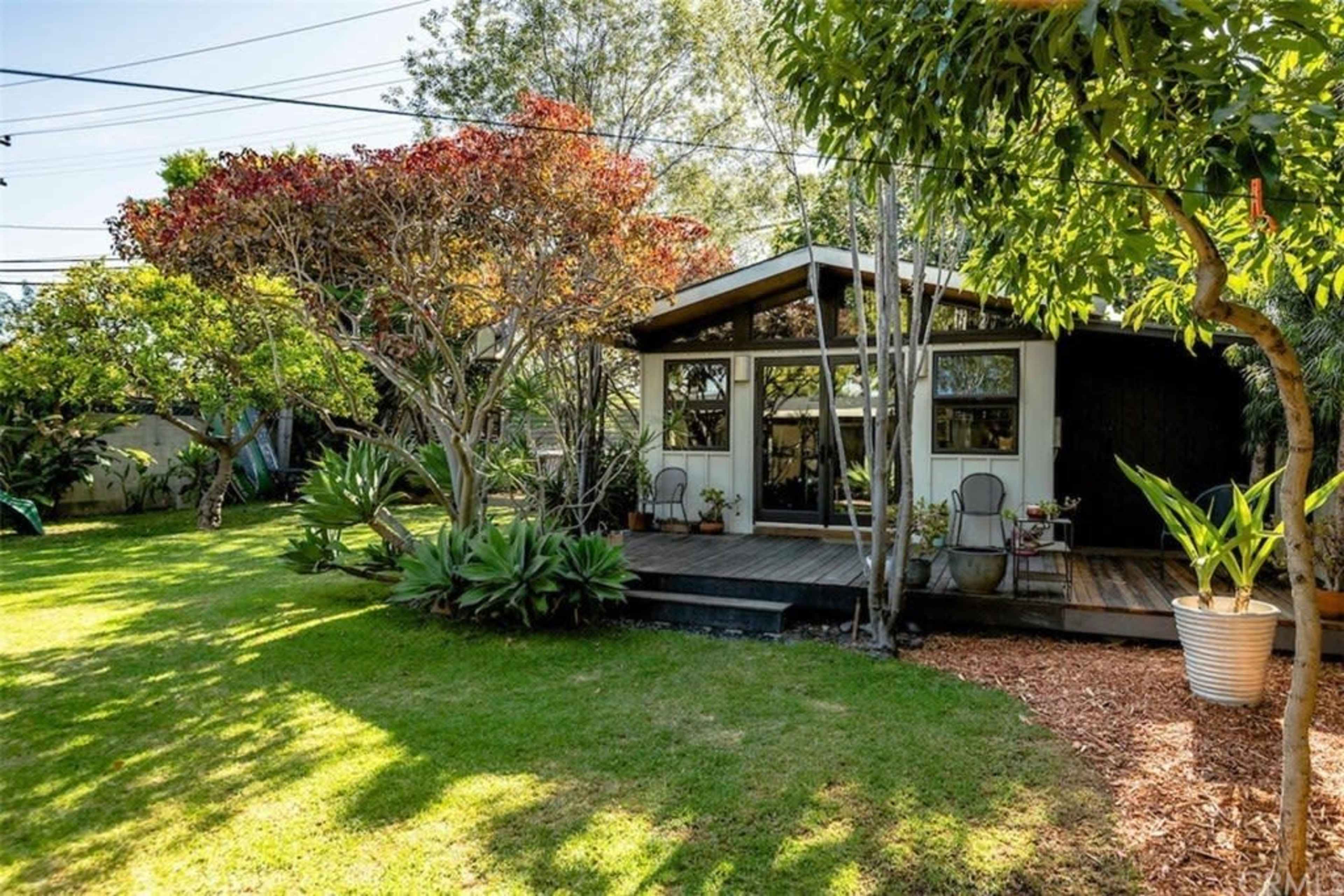 A small house with large windows is nestled among greenery and garden features in a backyard setting.