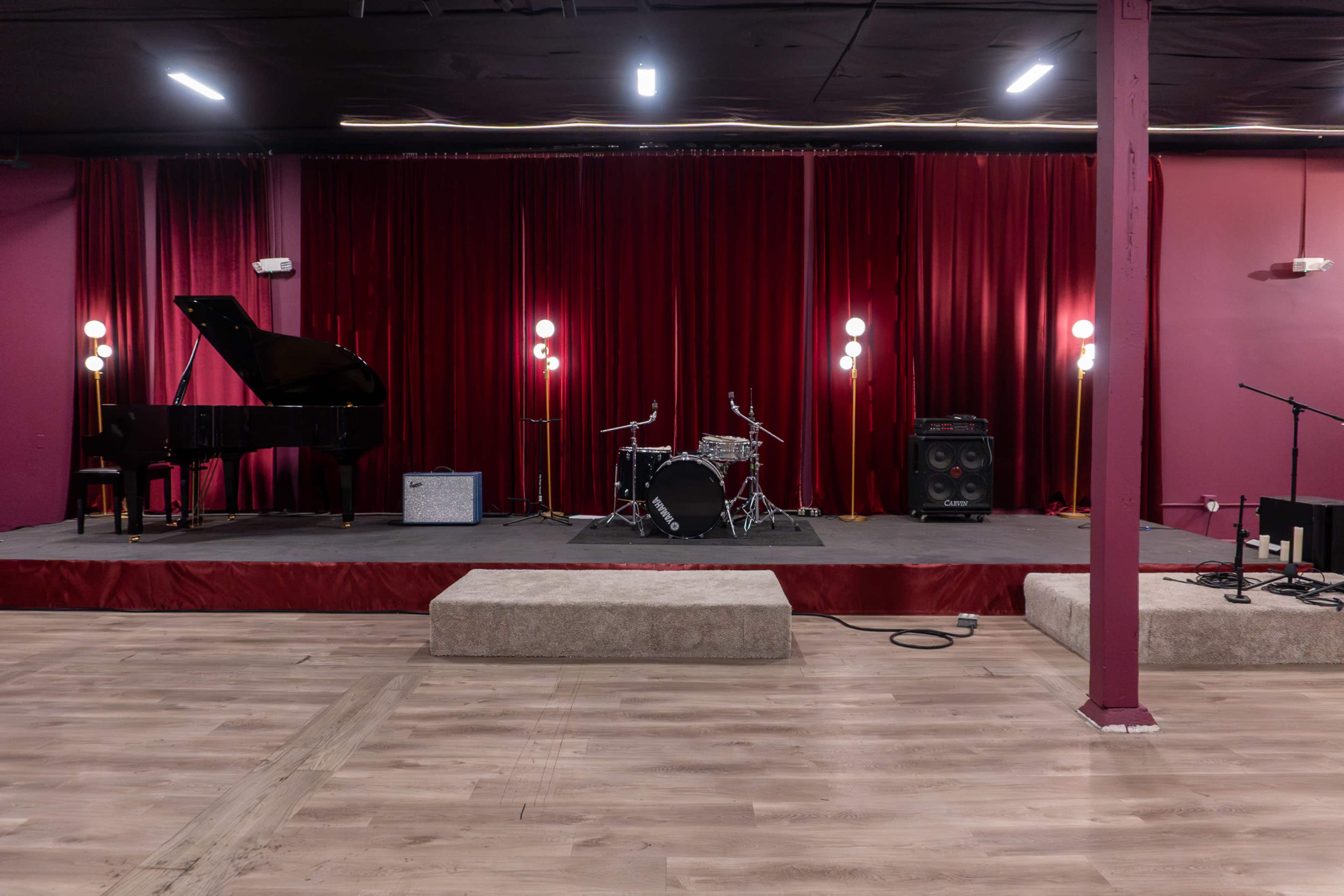 The image shows a stage with a grand piano, a drum set, amplifiers, and soft lighting in front of a red velvet backdrop.