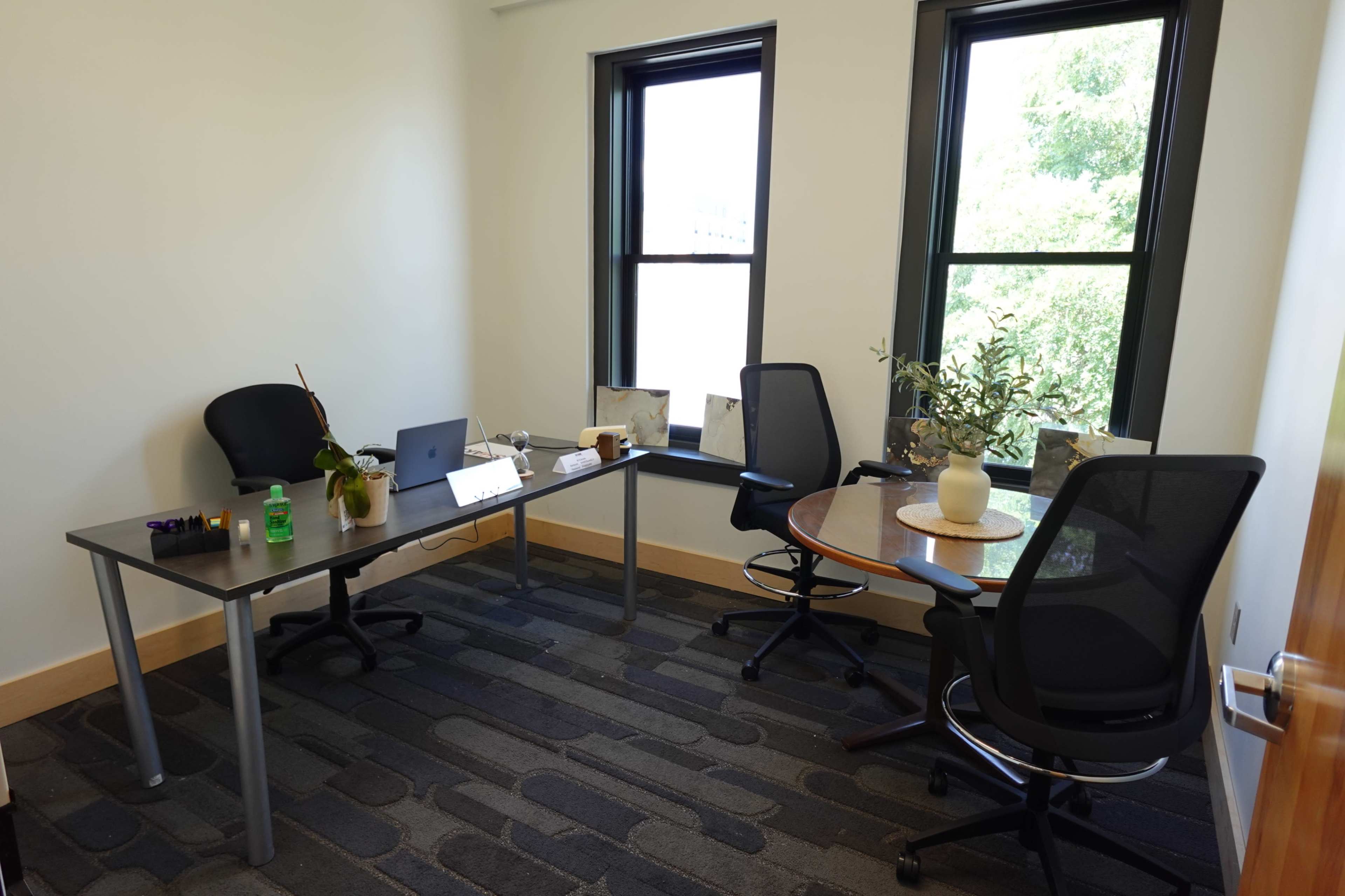 The image shows a small office space featuring a desk with a laptop, a plant, and two chairs, along with large windows allowing natural light to enter.