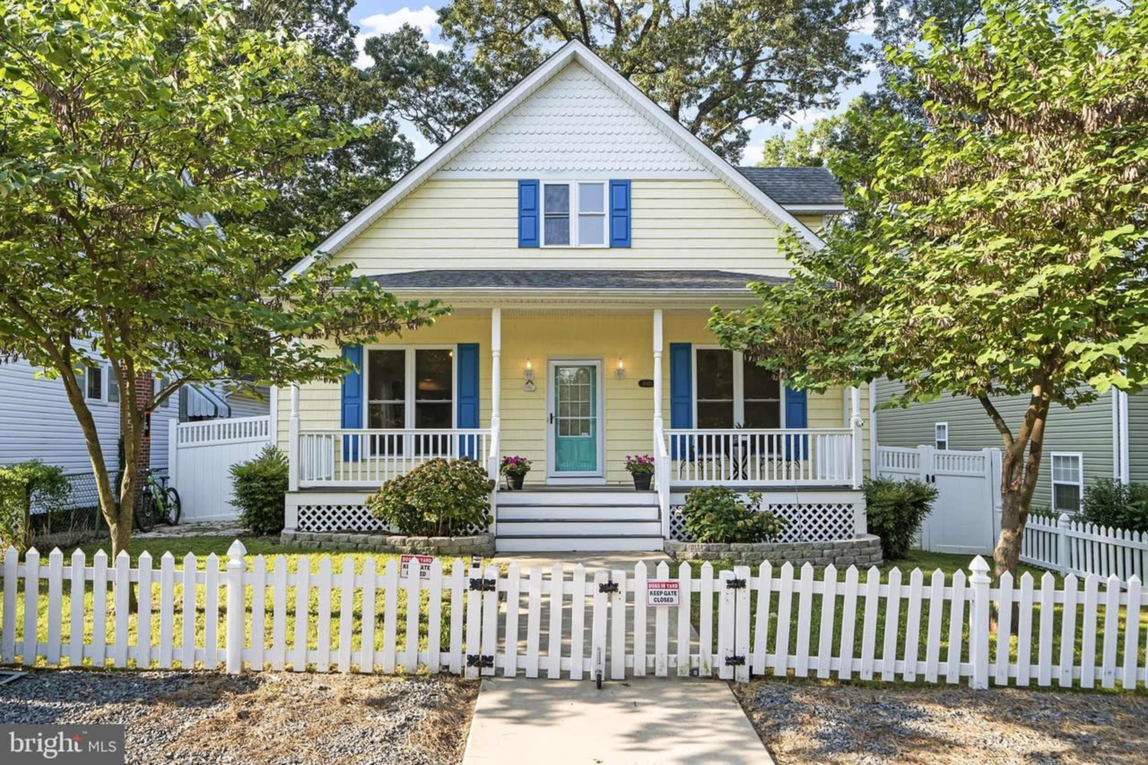 A yellow suburban house with a white picket fence, flanked by trees and featuring a front porch with blue shutters.