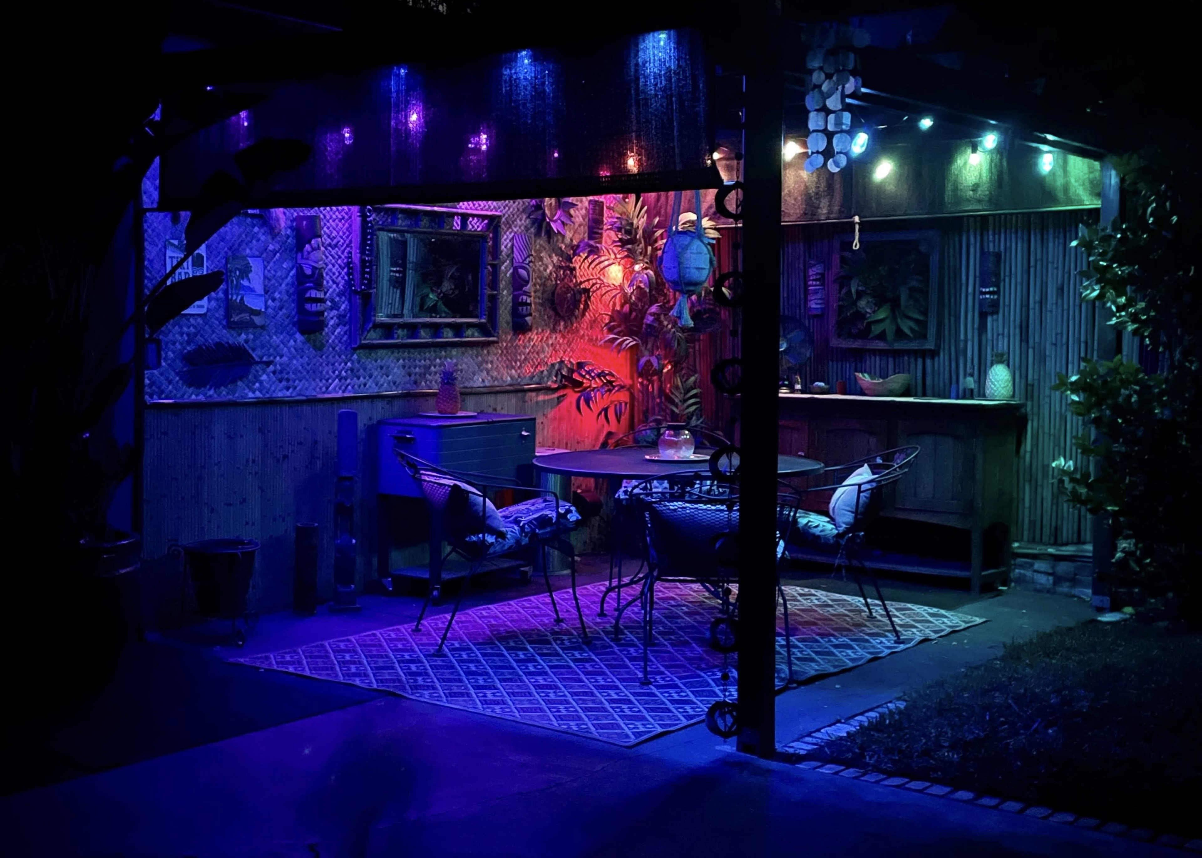 A patio is illuminated with colorful LED lights, featuring plants, a dining set, and decorative wall hangings.