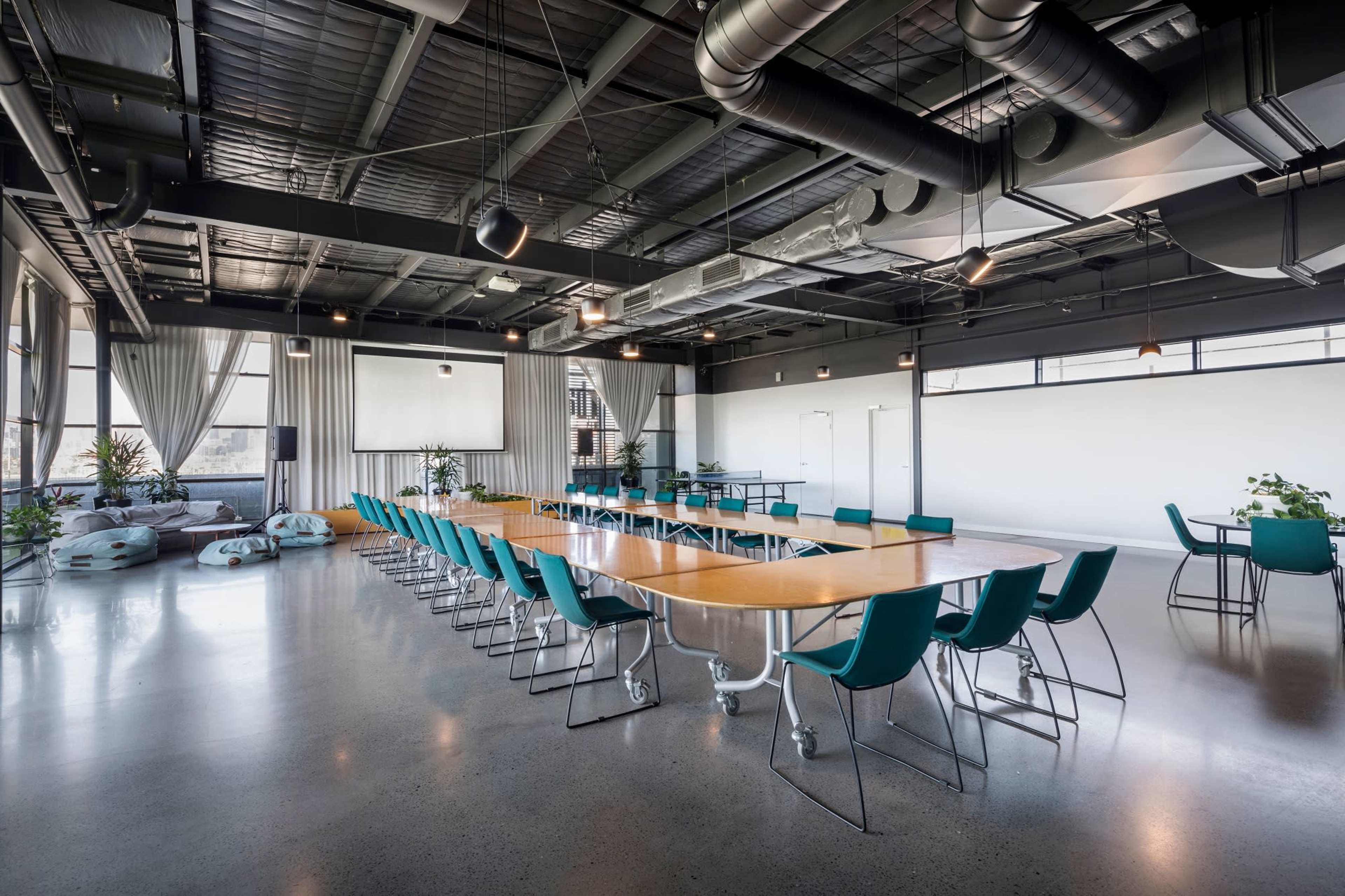 A large, modern conference room is set up with a long table and teal chairs, featuring industrial-style ceilings and large windows.