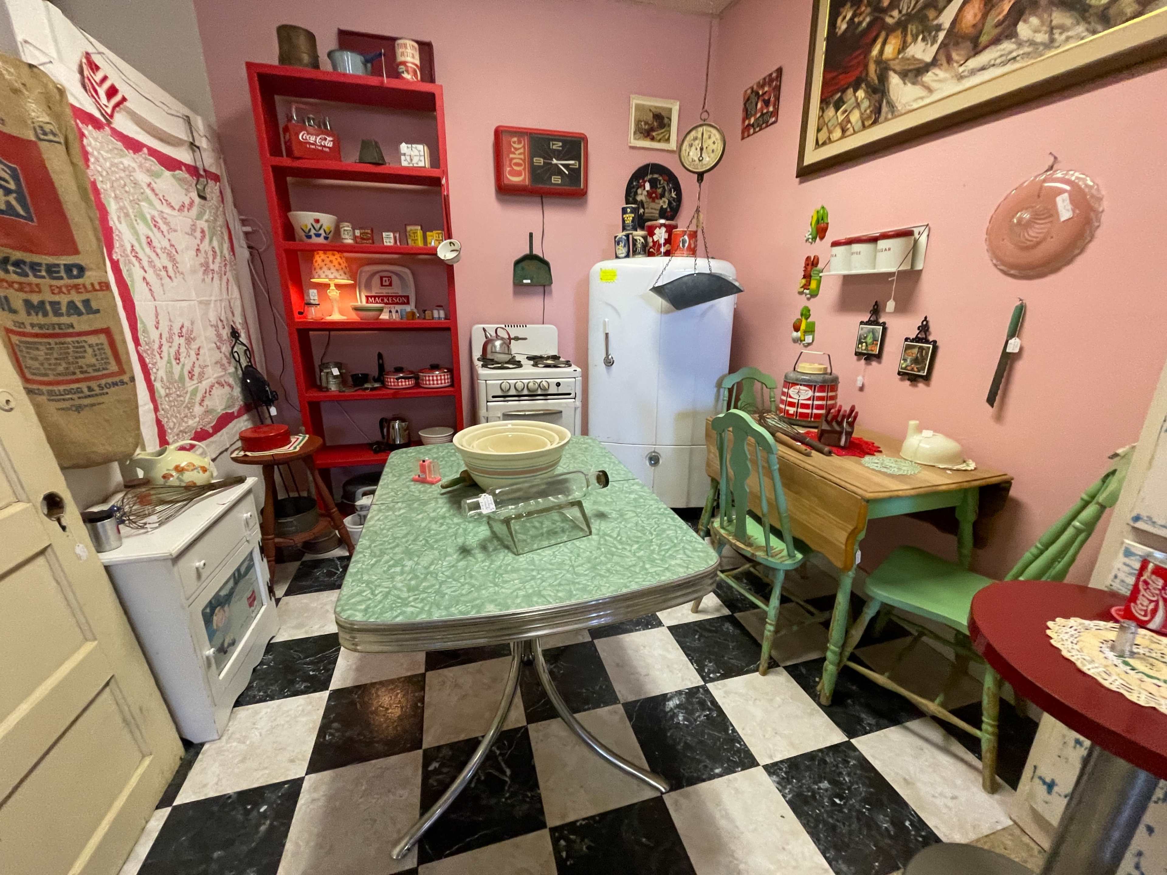 The image shows a retro kitchen with a checkered floor, a green table, vintage appliances, and various decorations on the walls and shelves.